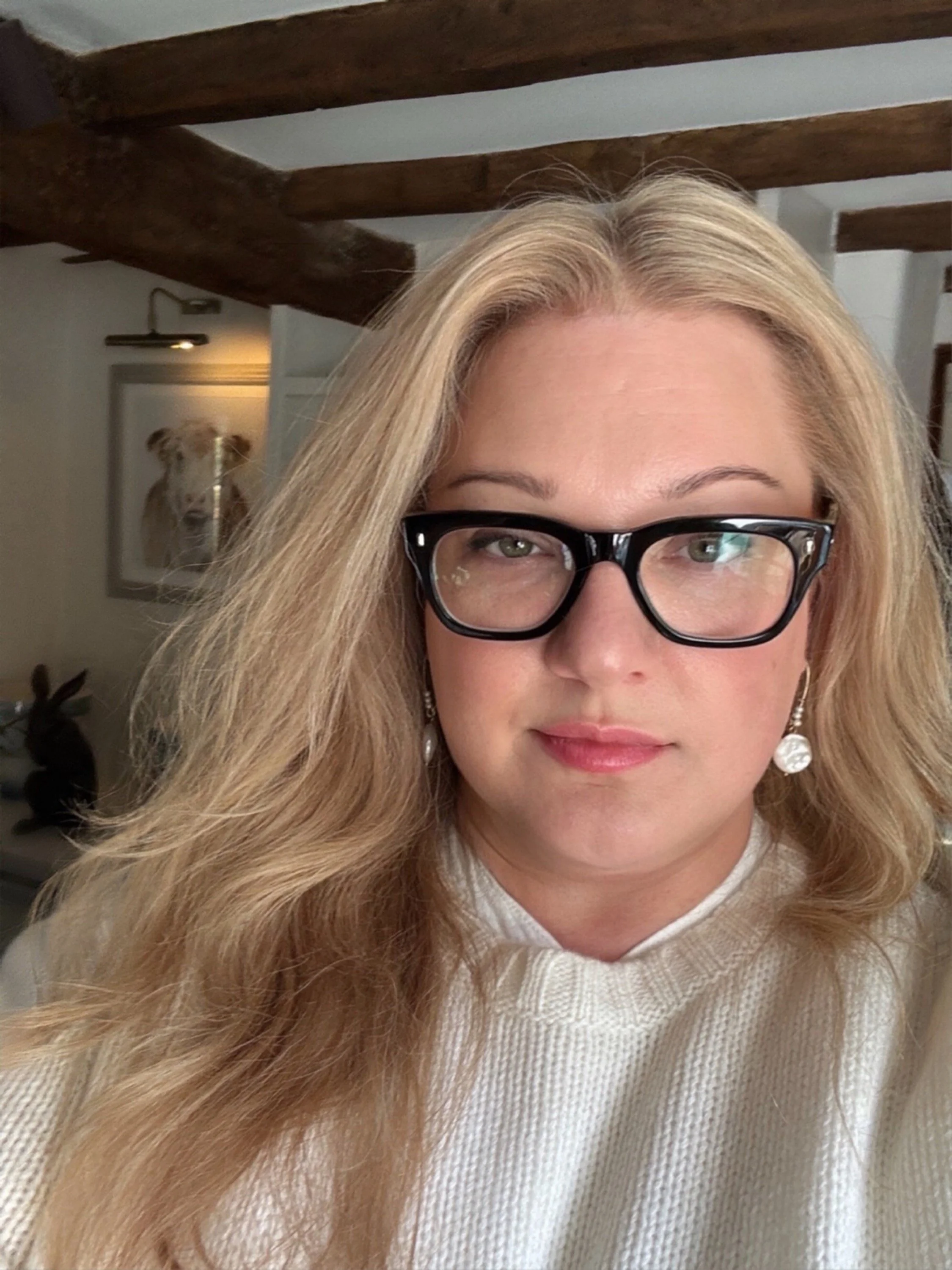 A close-up photo of a woman with blonde hair and black glasses, wearing a white knit sweater, with a dog picture in the background.