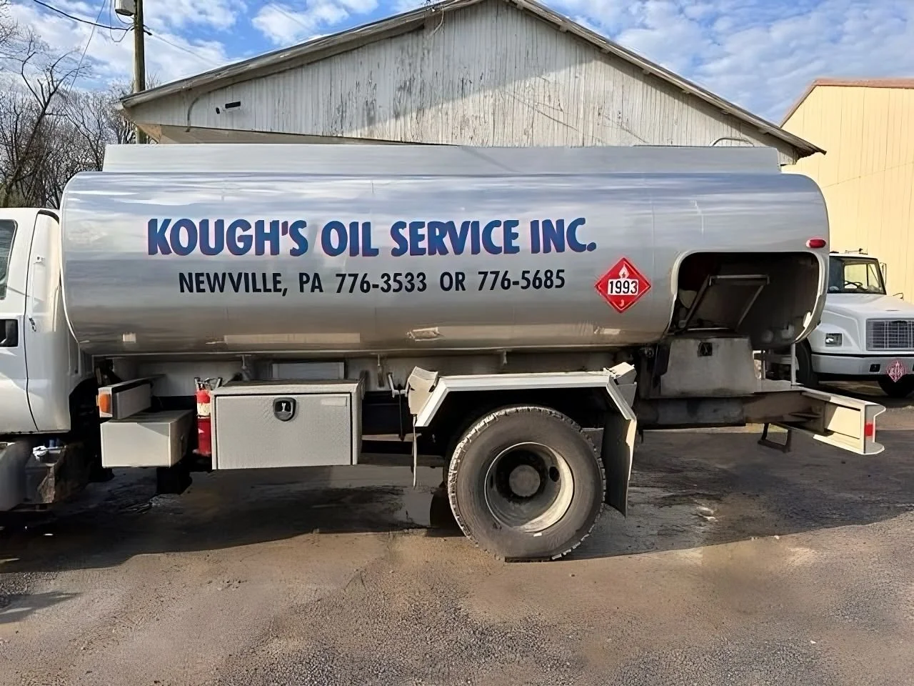 A fuel tanker truck labeled "Kough's Oil Service Inc.", located in Newville, Pennsylvania, with contact numbers 776-3533 and 776-5685. The tanker is gray with a red hazard label marked 1993. The tank is parked outdoors on a dirt surface with buildings and other vehicles in the background.