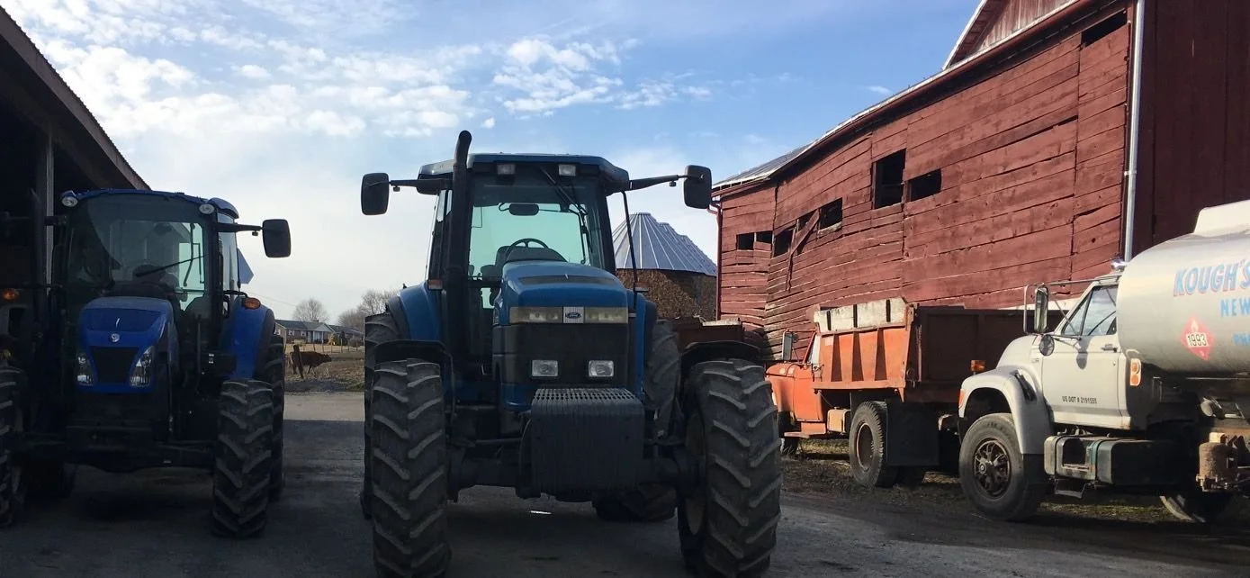 Three blue tractors, a red barn with damaged siding, and a white tanker truck parked on a farm driveway, with a partly cloudy sky in the background.