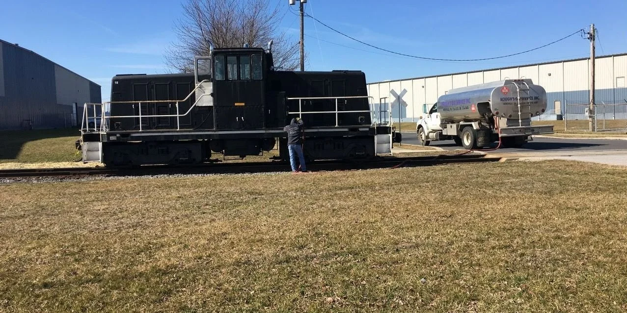 A small black train engine on a railroad track with a person inspecting it, and a fuel truck connected to the train on the side.