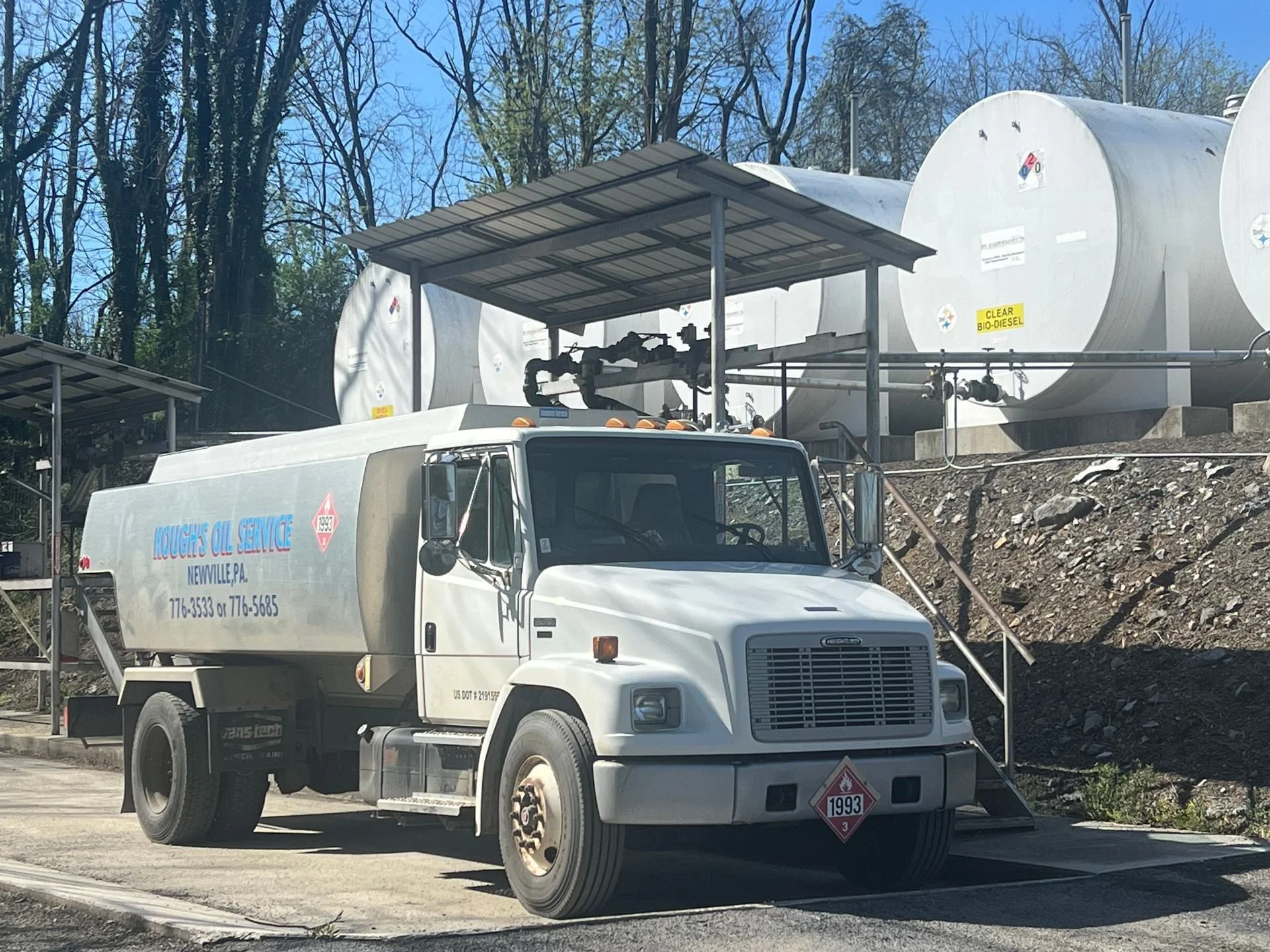 White oil service truck with company name and contact info, parked outside with large bio-diesel storage tanks in the background, trees, and a blue sky.