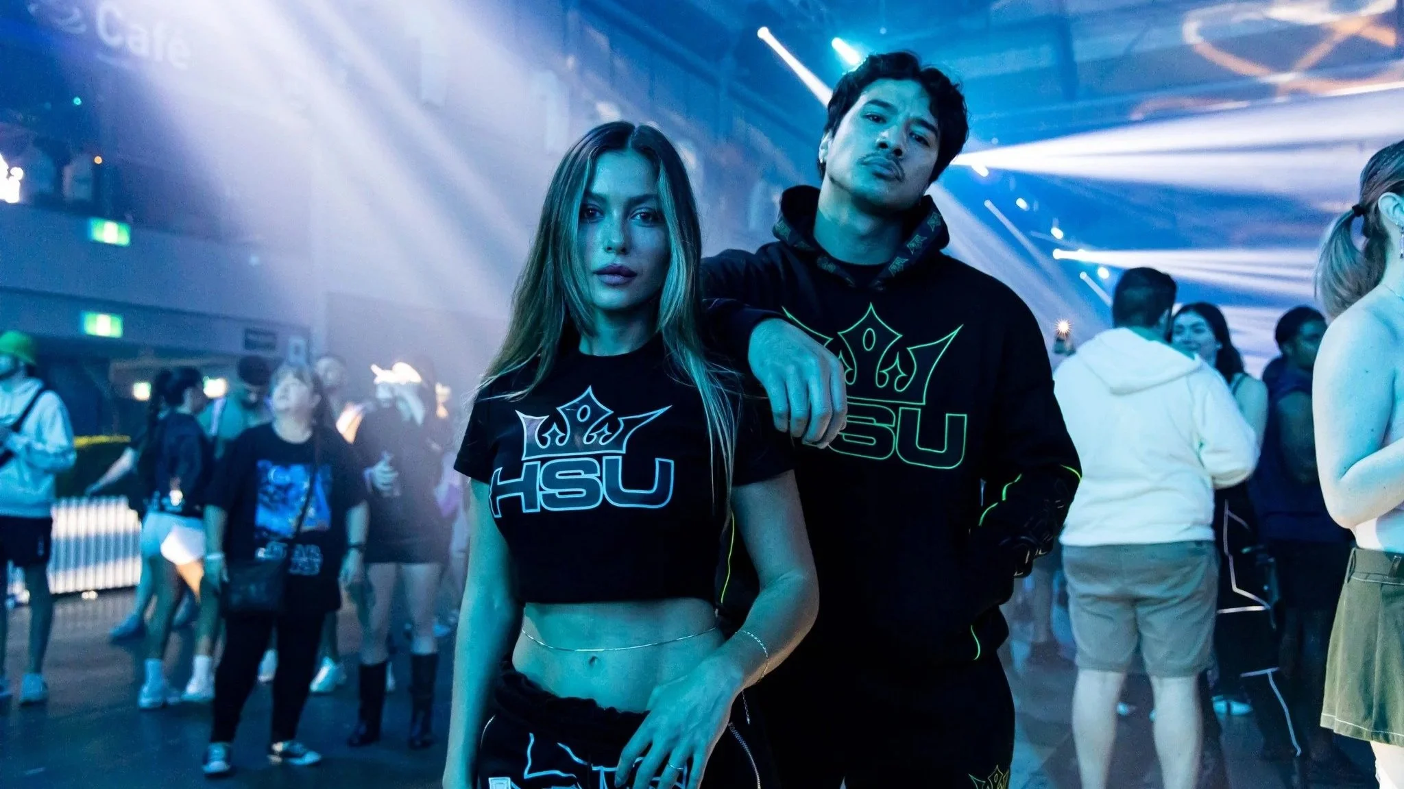 Two people standing in a crowded indoor event, wearing black clothing with 'HSU' and a crown logo, illuminated by blue and white stage lights, with many other attendees in the background.