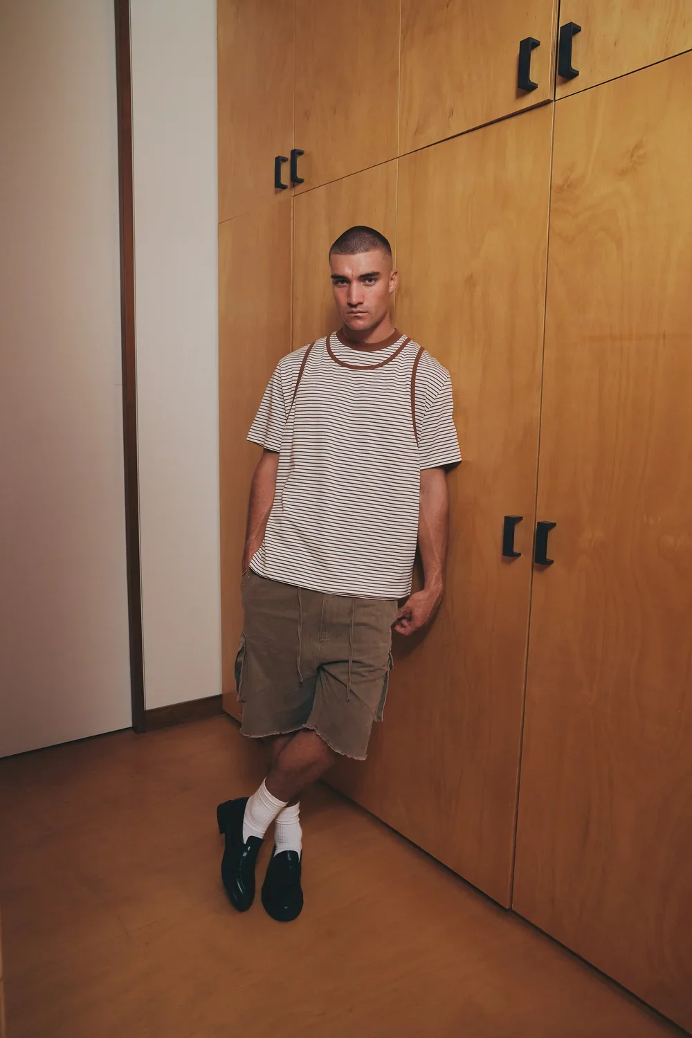 A young man with a buzz cut hairstyle standing against a wooden wall, wearing a striped t-shirt, khaki cargo shorts, white socks, and black shoes, with one hand in his pocket and the other hanging by his side.