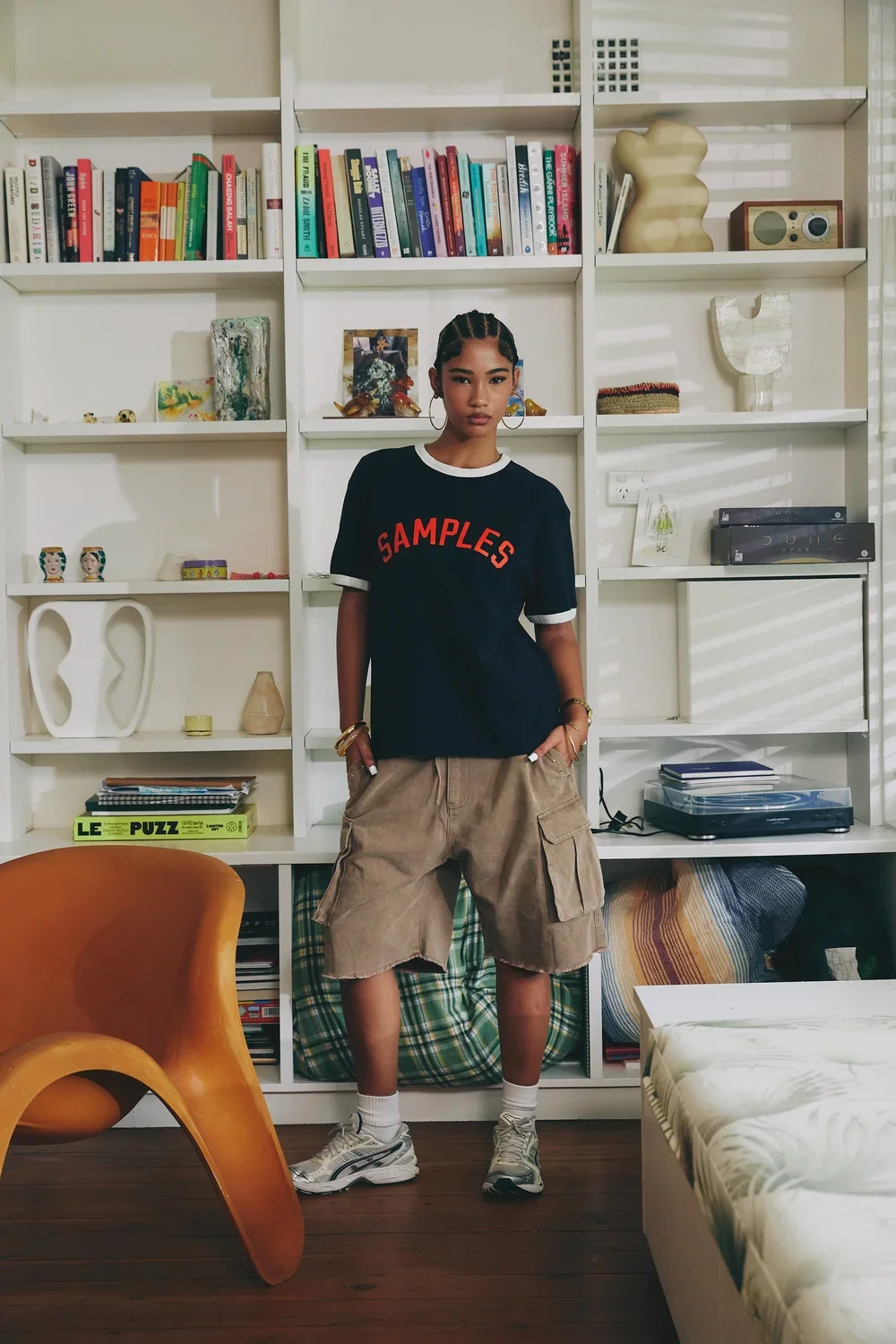 A young woman with braided hair and hoop earrings stands in a living room in front of a white bookshelf filled with books, art, and decorative objects. She is wearing a black t-shirt with red lettering that says 'SAMPLES', beige cargo shorts, white sneakers, and white socks. She has jewelry on her wrists and hands and stands with her hands in her pockets.
