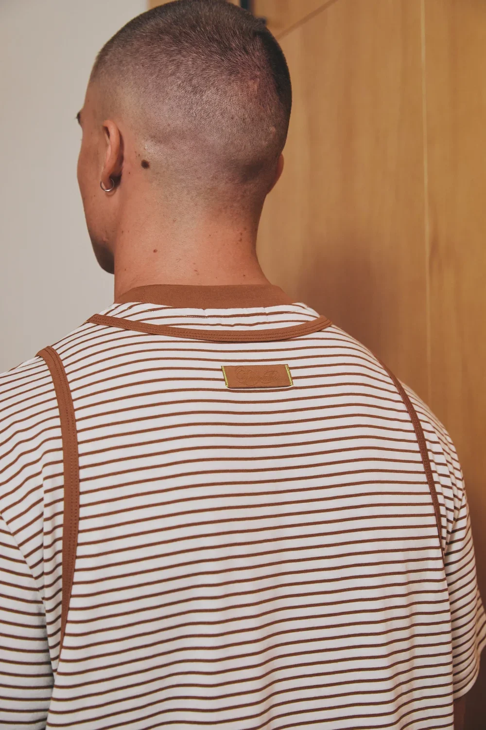 Back of a person wearing a white and brown striped shirt, with a close-cropped hairstyle, a small hoop earring, standing in front of a wooden door.