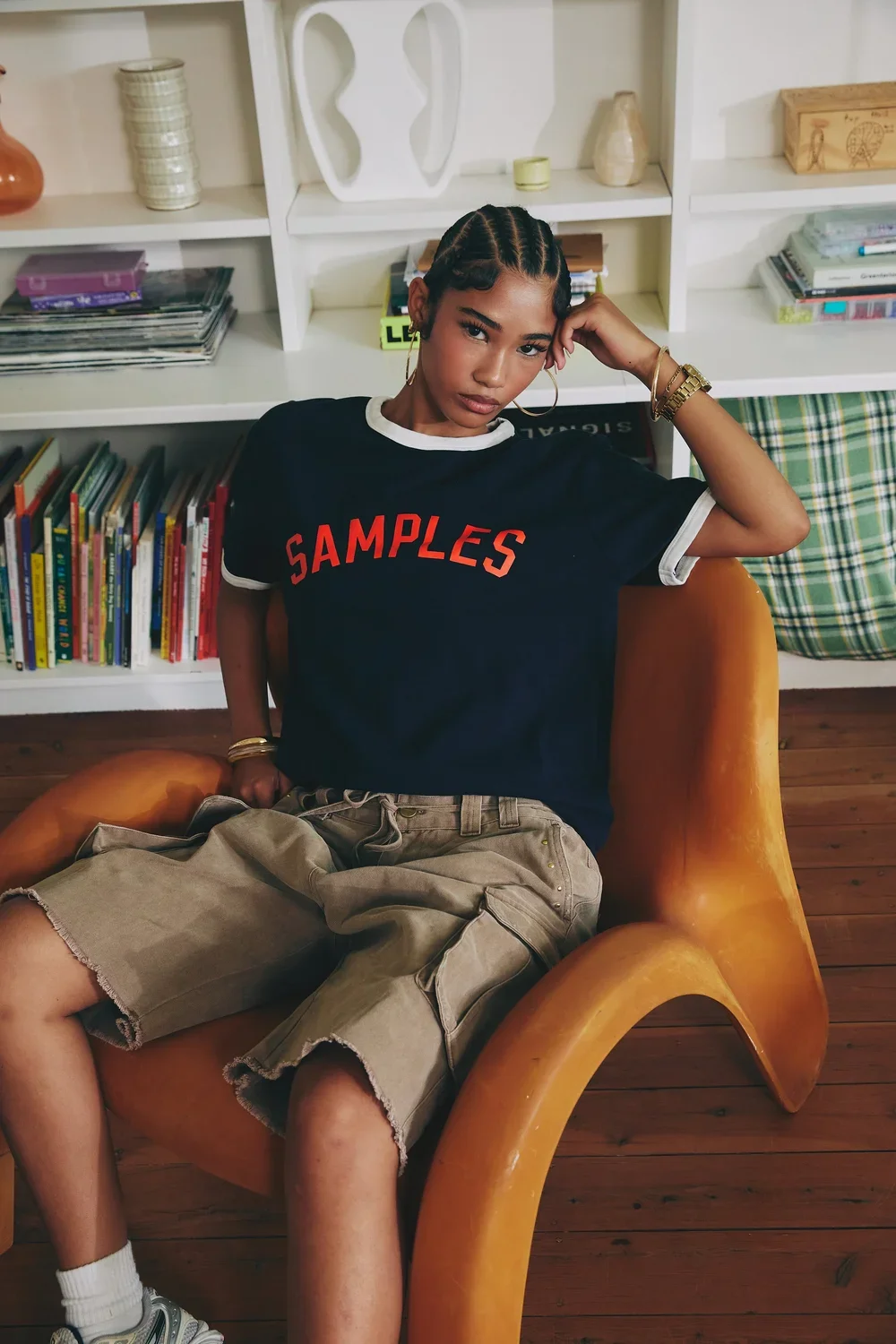 A young woman with braided hair, wearing a black t-shirt with red lettering that says 'SAMPLES', tan shorts, and multiple gold jewelry pieces, sitting on an orange chair in front of a white bookshelf filled with books and decorative items.