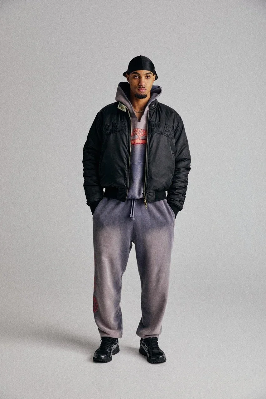 A young man is standing against a plain gray background. He is wearing a black bomber jacket over a gray hoodie, gray sweatpants, black sneakers, and a black durag.