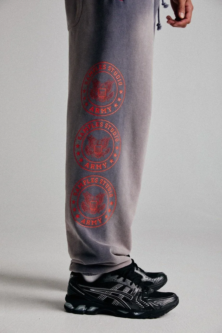 Person wearing grey sweatpants with red printed army and studio logos, black athletic shoes, and standing against a plain background.