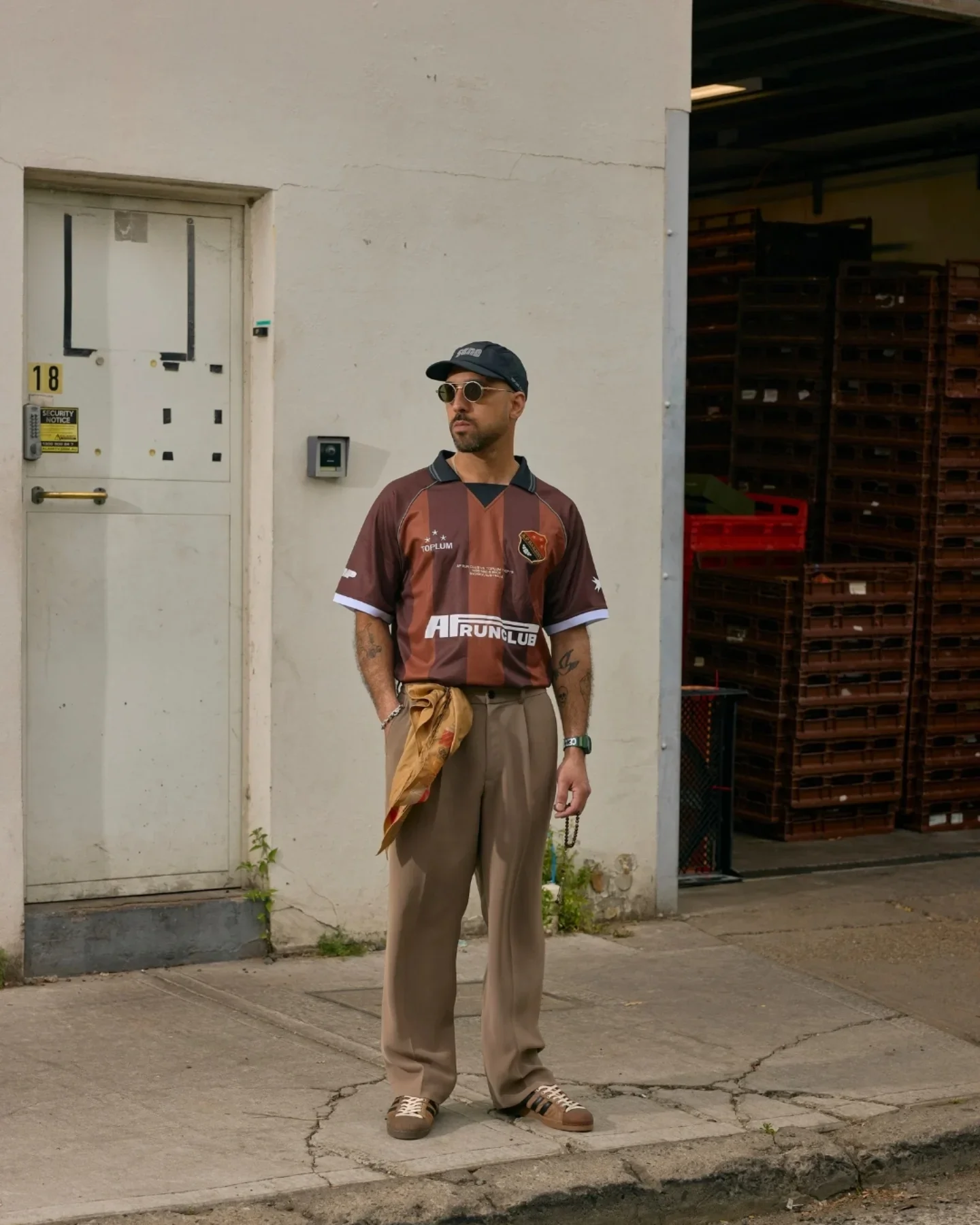 A man standing outside in front of a white building wall. He is wearing sunglasses, a black cap, a maroon sports jersey with various logos, loose beige pants, and brown sneakers. He has tattoos on his arms and is holding a small object and an item of clothing in his left hand. To the right, there are stacked brown crates or pallets.