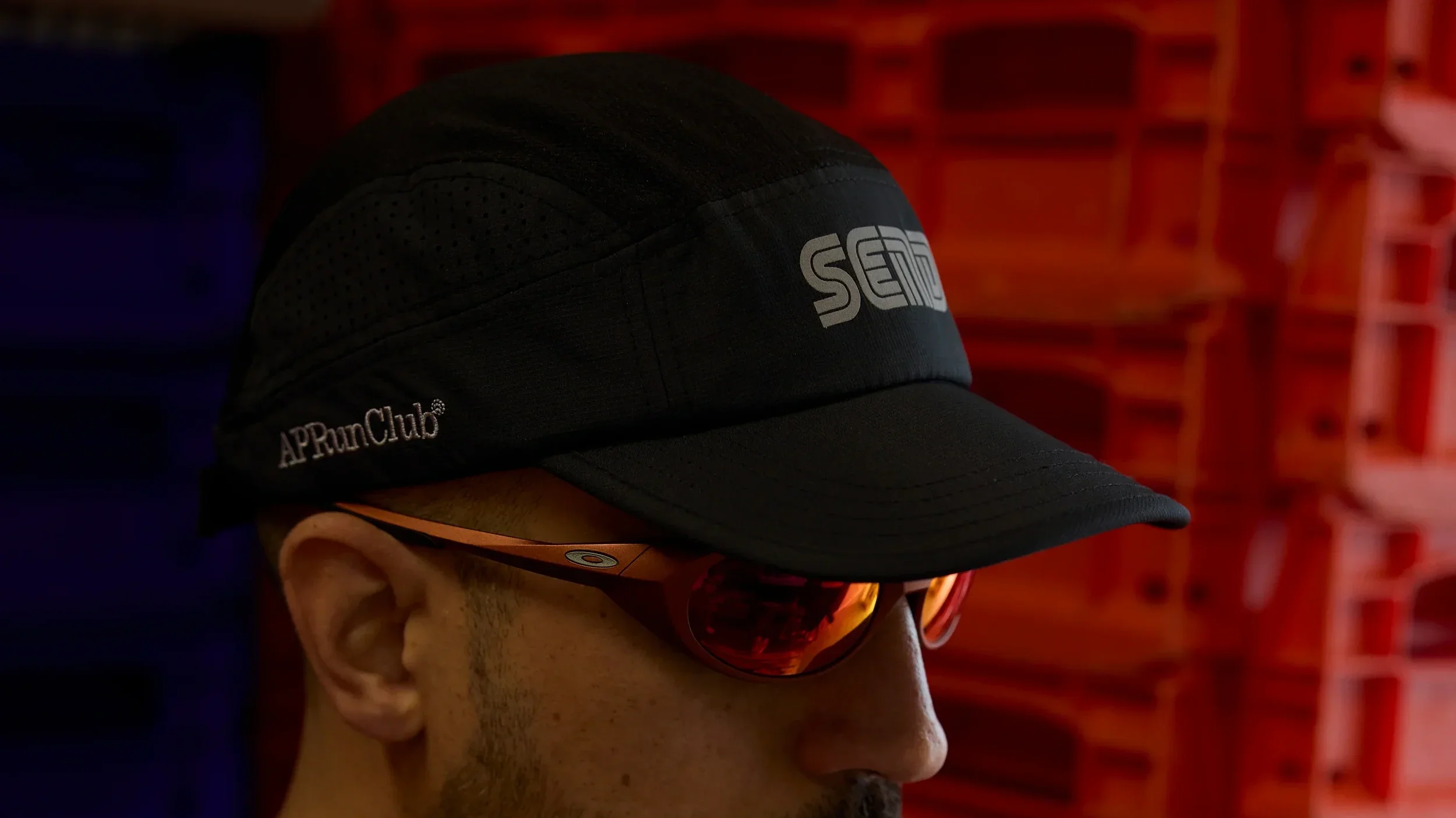Side profile of a person wearing a black running cap with 'APRunClub' logo, orange sunglasses, and a sports shirt, with red storage bins in the background.