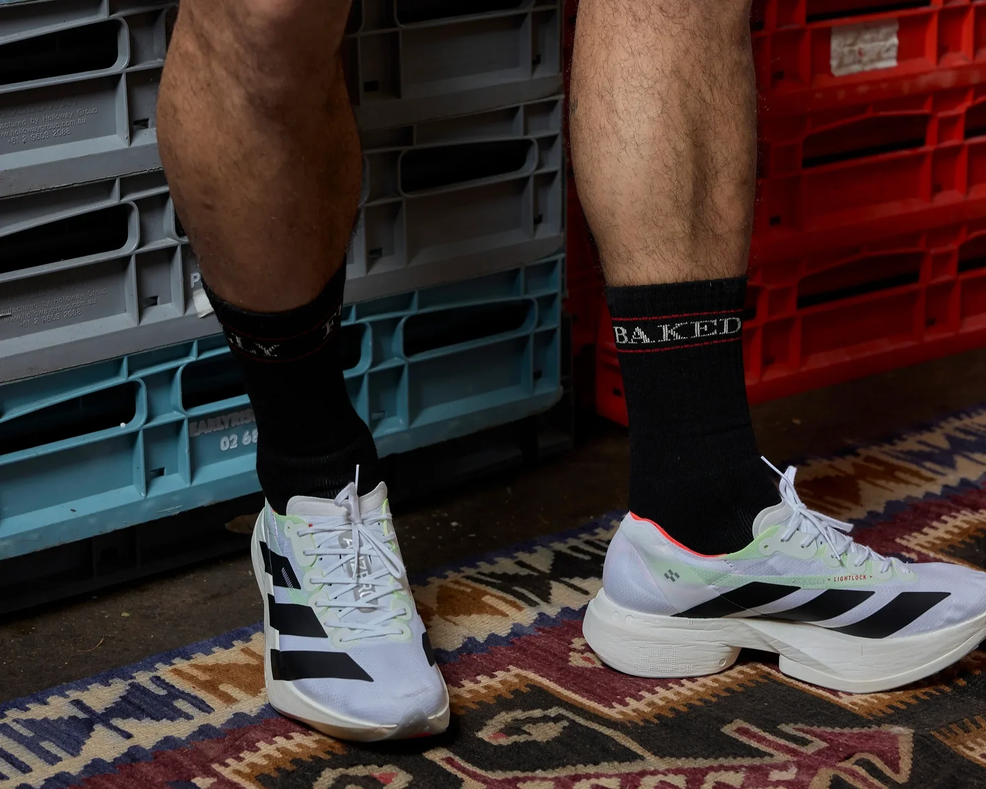 Person wearing white and black athletic shoes, black socks, and white sports shoes, standing on a patterned rug, with grey and red storage bins in the background.