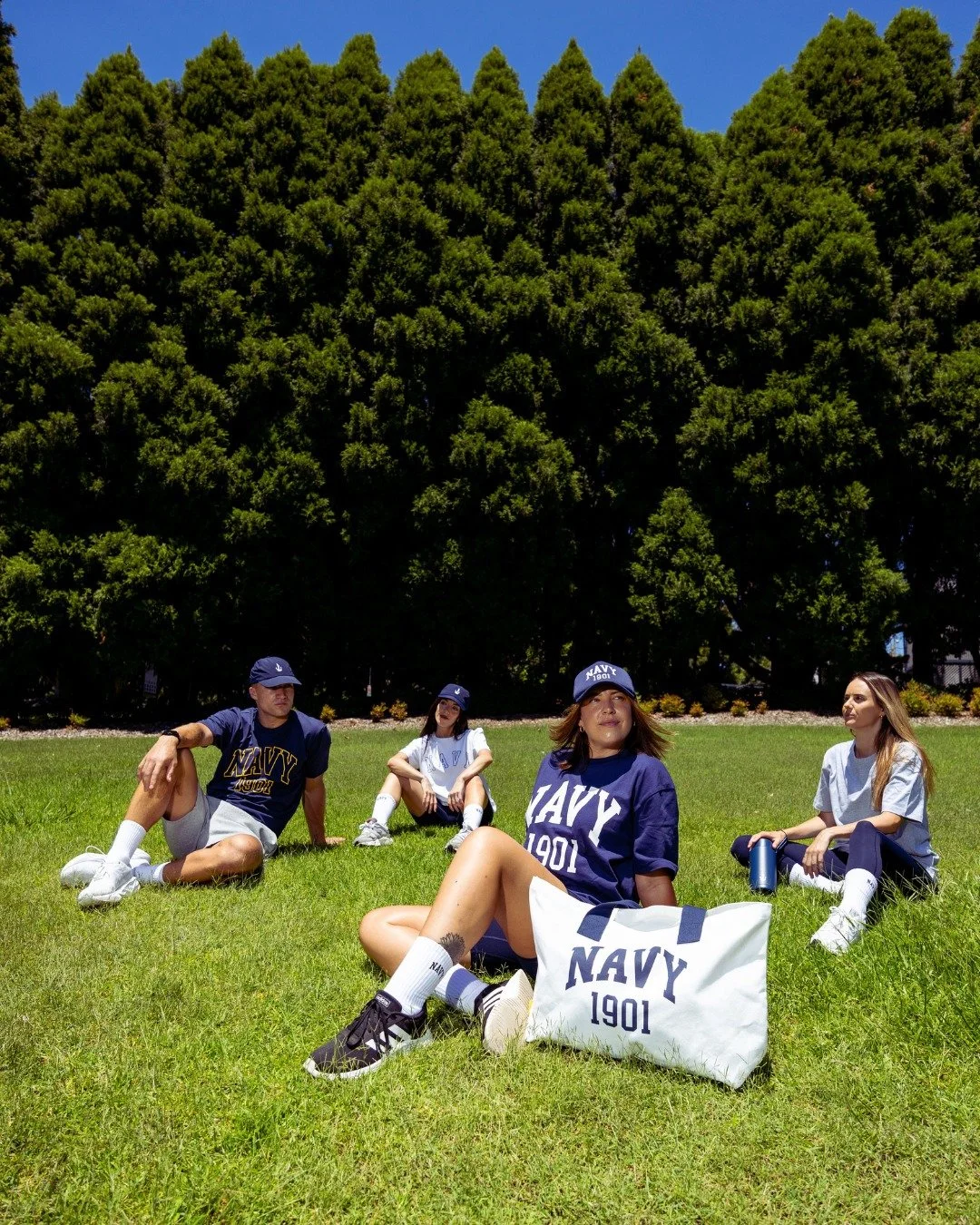 Four young women and one young man outdoors on a grassy field, wearing navy-themed clothing and caps, seated near a large green tree under a clear blue sky.