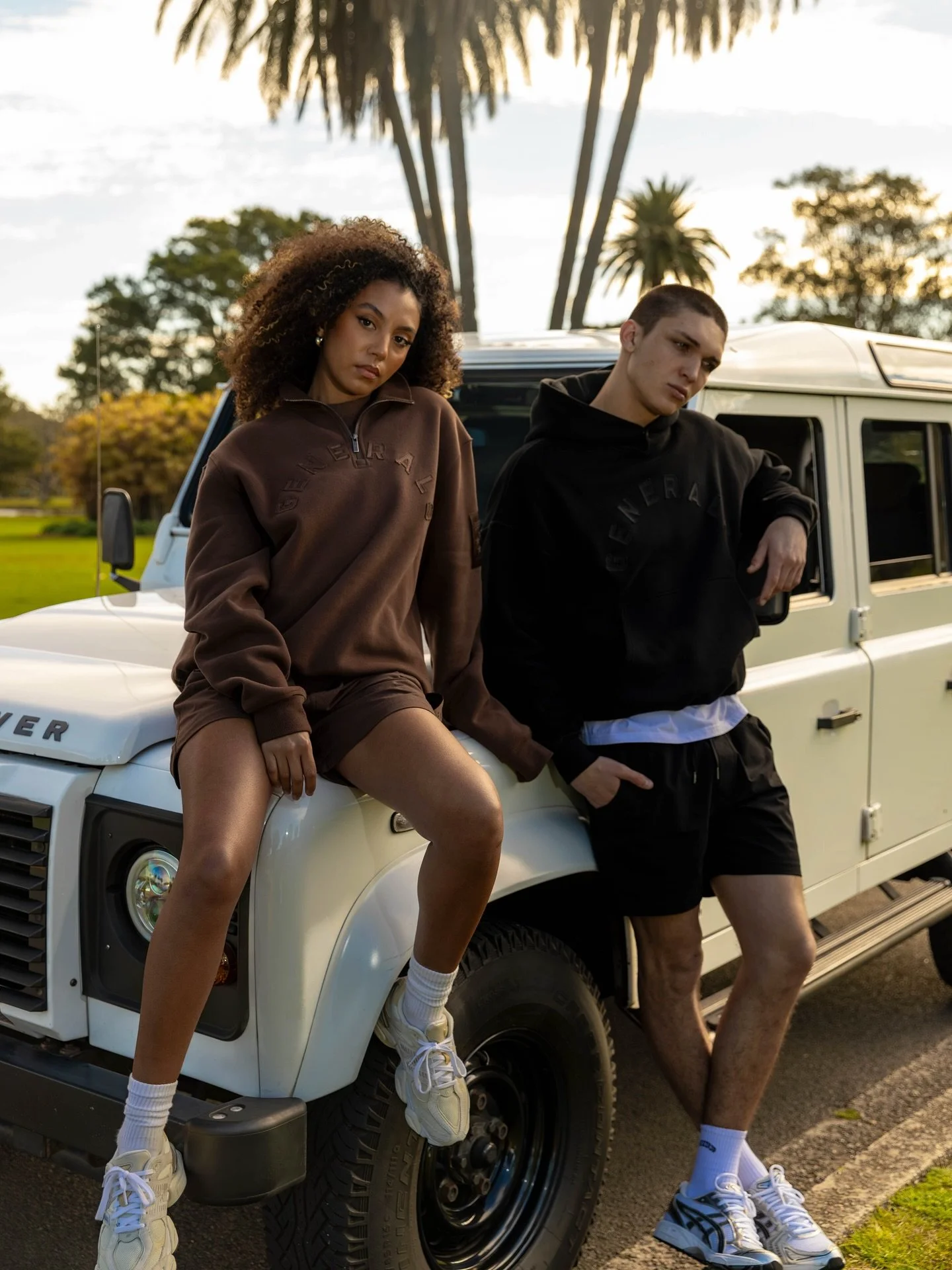 Two young adults, a woman with curly hair and a man with short hair, pose casually on a white Land Rover parked outdoors with trees and palm trees in the background. They are dressed in casual streetwear and sneakers, with the woman sitting on the front of the vehicle and the man leaning against it.