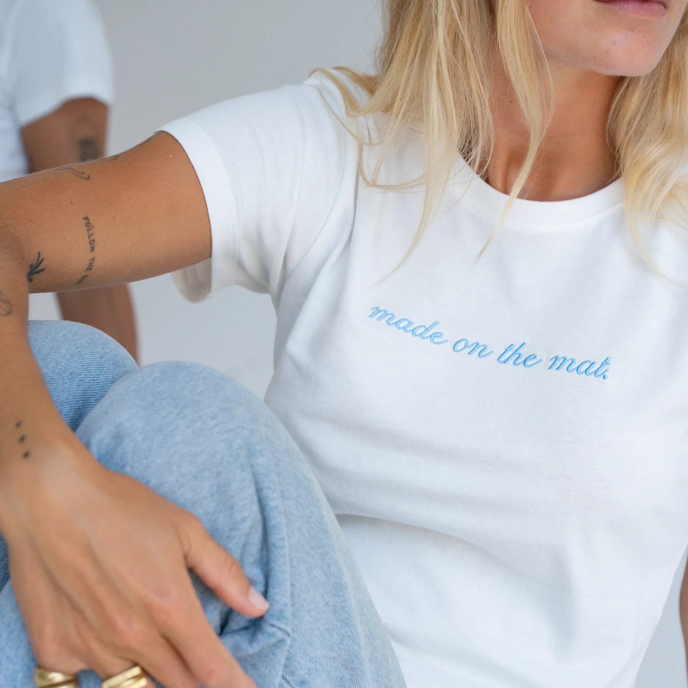 A person with blonde hair wearing a white shirt, holding a grayish-blue object, with visible tattoos on their arm and wearing rings on their fingers.