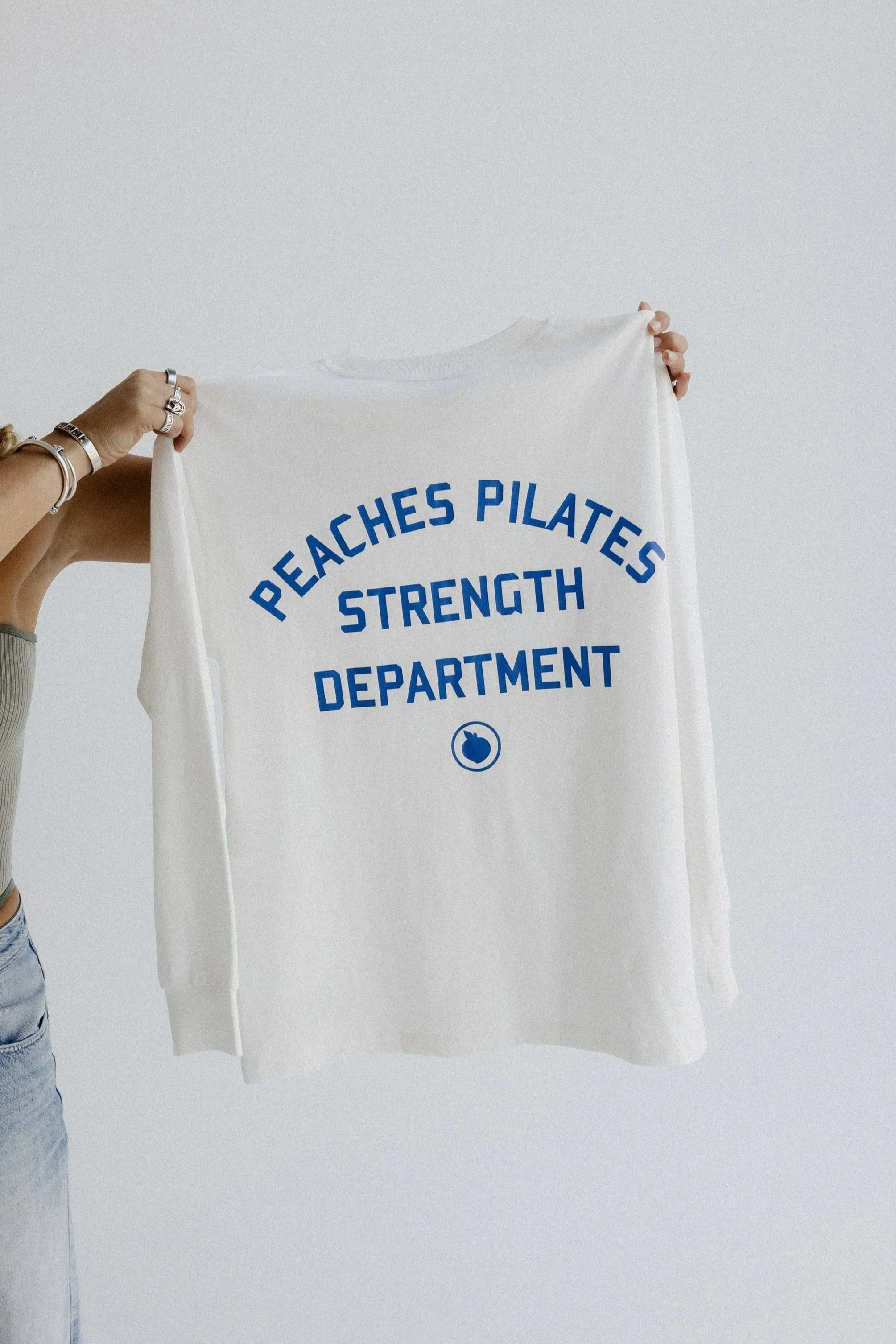 Person holding a white towel or cloth with blue printed text that reads 'PEACHES PILATES STRENGTH DEPARTMENT' against a plain light-colored background.