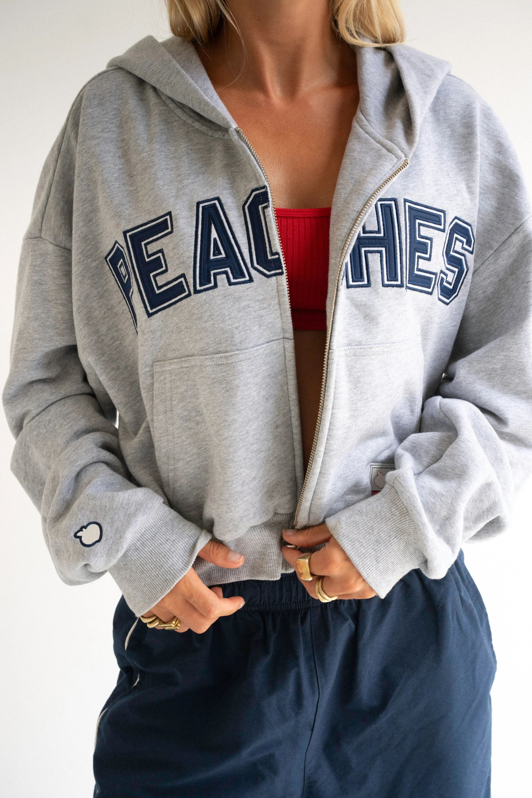 Person wearing a gray hoodie with 'PEACE' written across the front, a red sports bra underneath, and navy blue pants. The person is adjusting their pants with their hands, which are adorned with multiple rings.