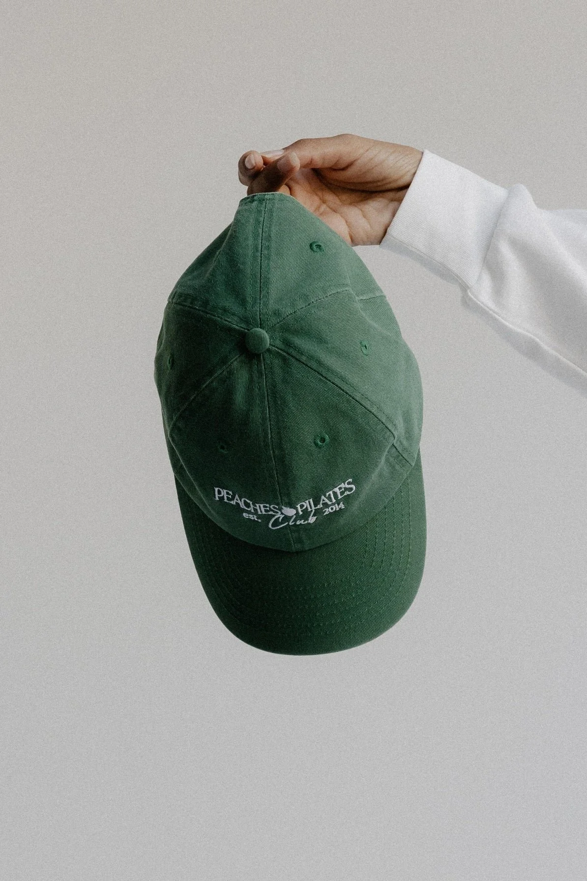 A green baseball cap with white embroidered text that reads 'Peaches Pilates Club est. 2014' is held by a person wearing a white long-sleeve shirt against a plain background.