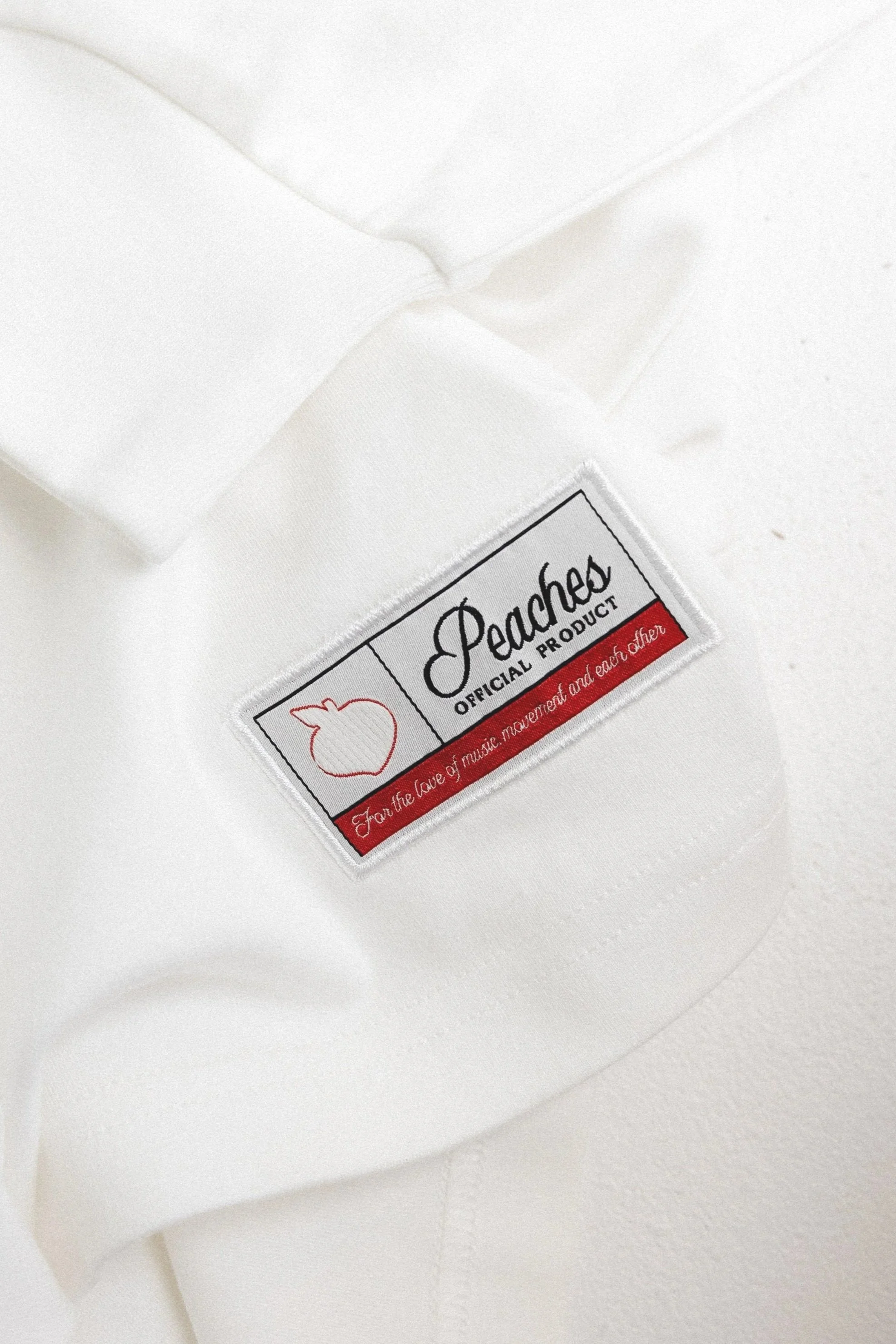 Close-up of a white garment with a label reading 'Peaches Official Product' with a peach logo and the slogan 'For the love of music, movement and each other' on a white background.
