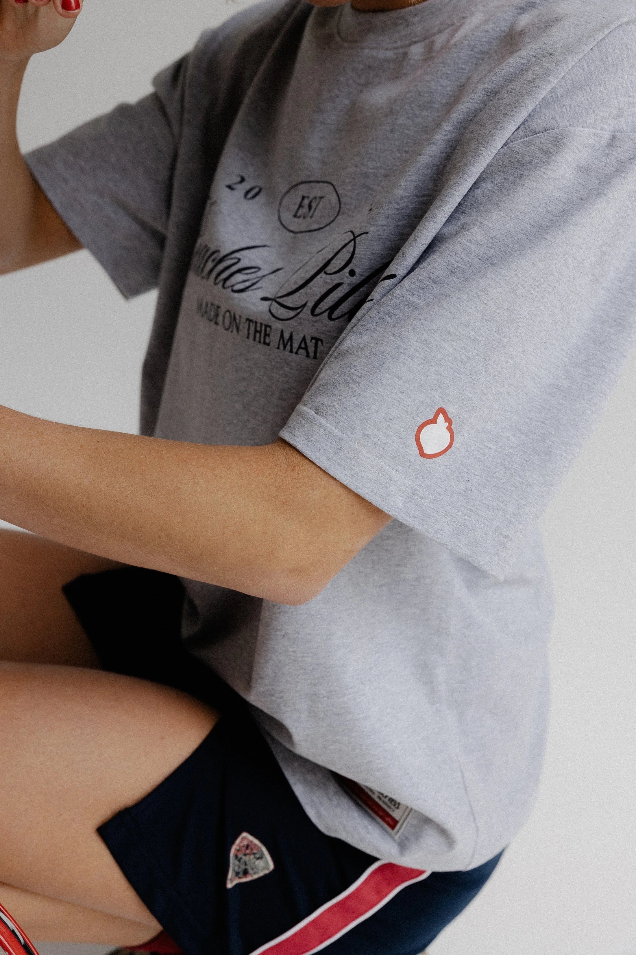 A person wearing a light gray T-shirt with black text and a small orange and white logo on the sleeve, and dark shorts with patches, standing against a plain background.