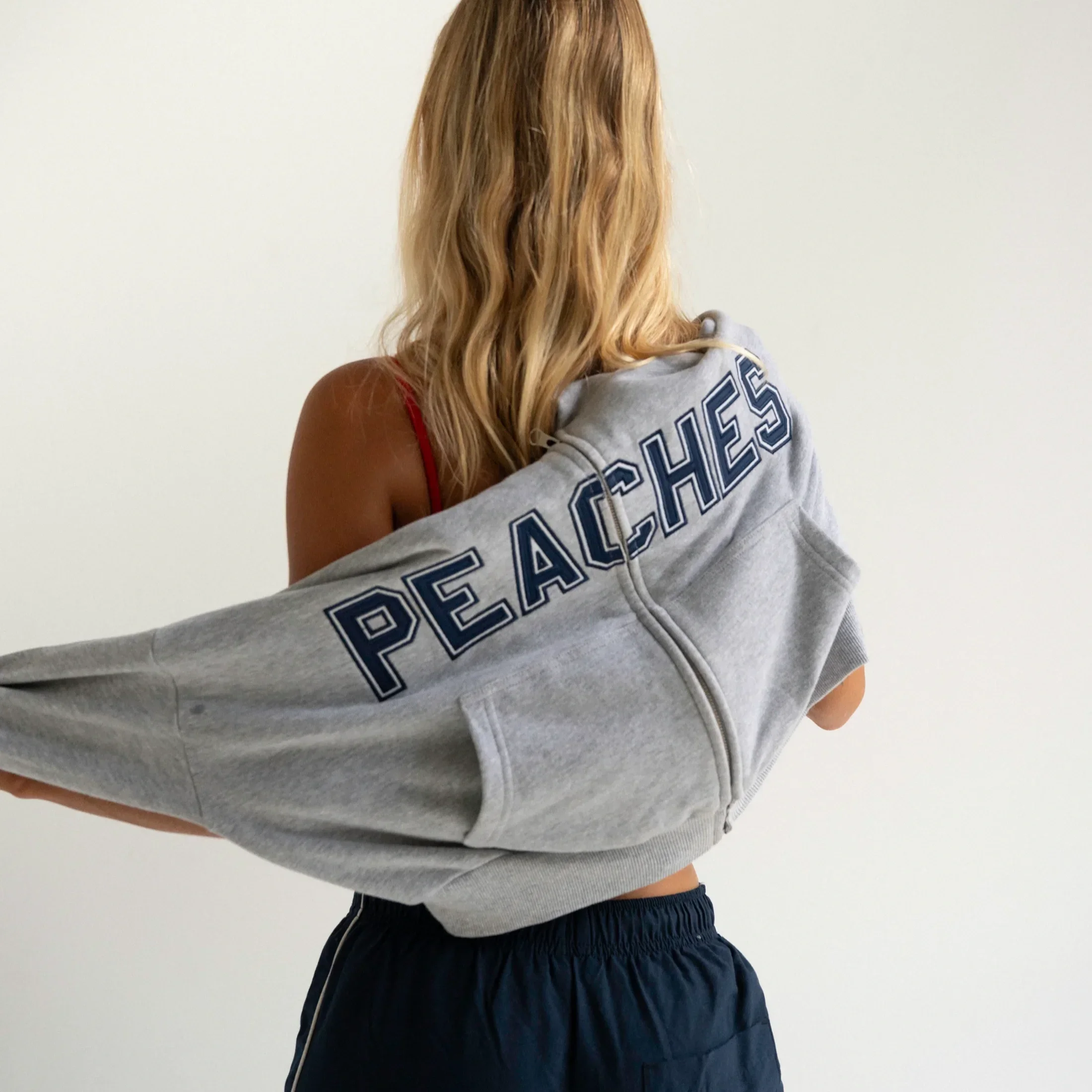 Back view of a woman with long blonde hair wearing a red tank top and holding a gray sweatshirt, standing against a plain white background.