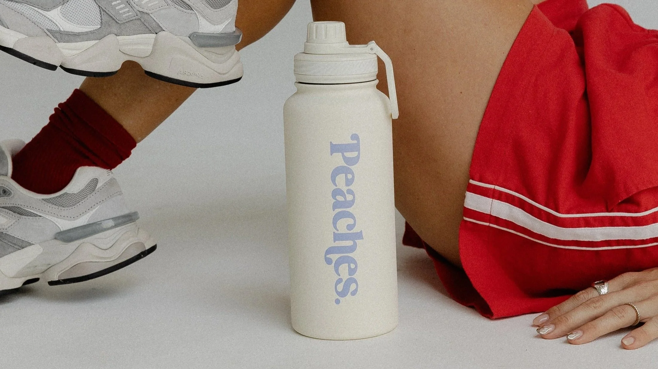 Close-up of a person's arm and hand resting on a white surface, wearing a red shirt with white stripe details, a wedding ring, and a ring on the index finger. Next to them is a white water bottle with 'space' written on it in light purple. There are also white sneakers with gray accents and red socks hanging from the person's leg.