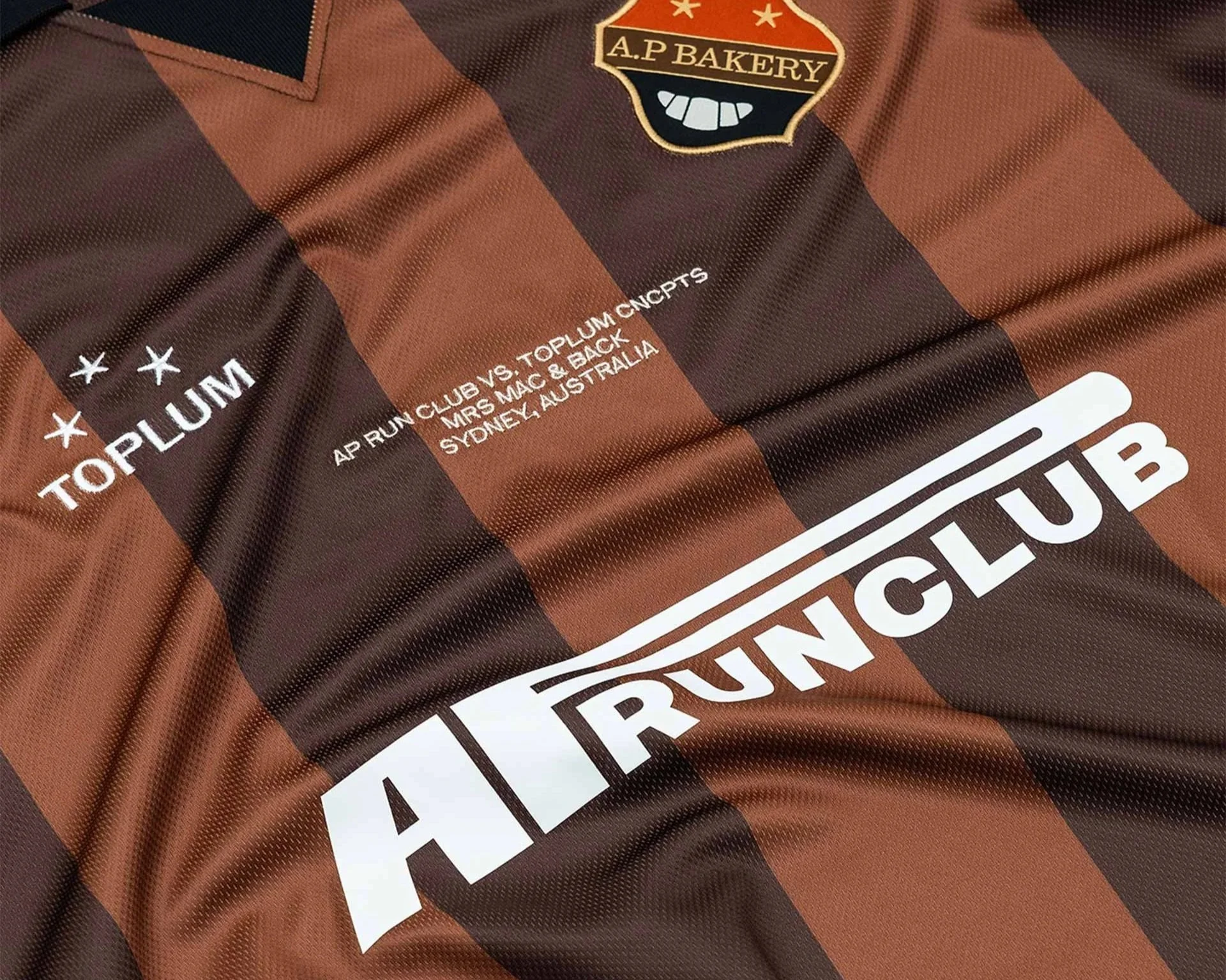 Close-up of a brown and black checkered athletic jersey with a patch reading 'A.P. Bakery' and an emblem of a croissant, along with text referencing a running club event between Newcastle and Toplum. The jersey also displays parts of other text in white.