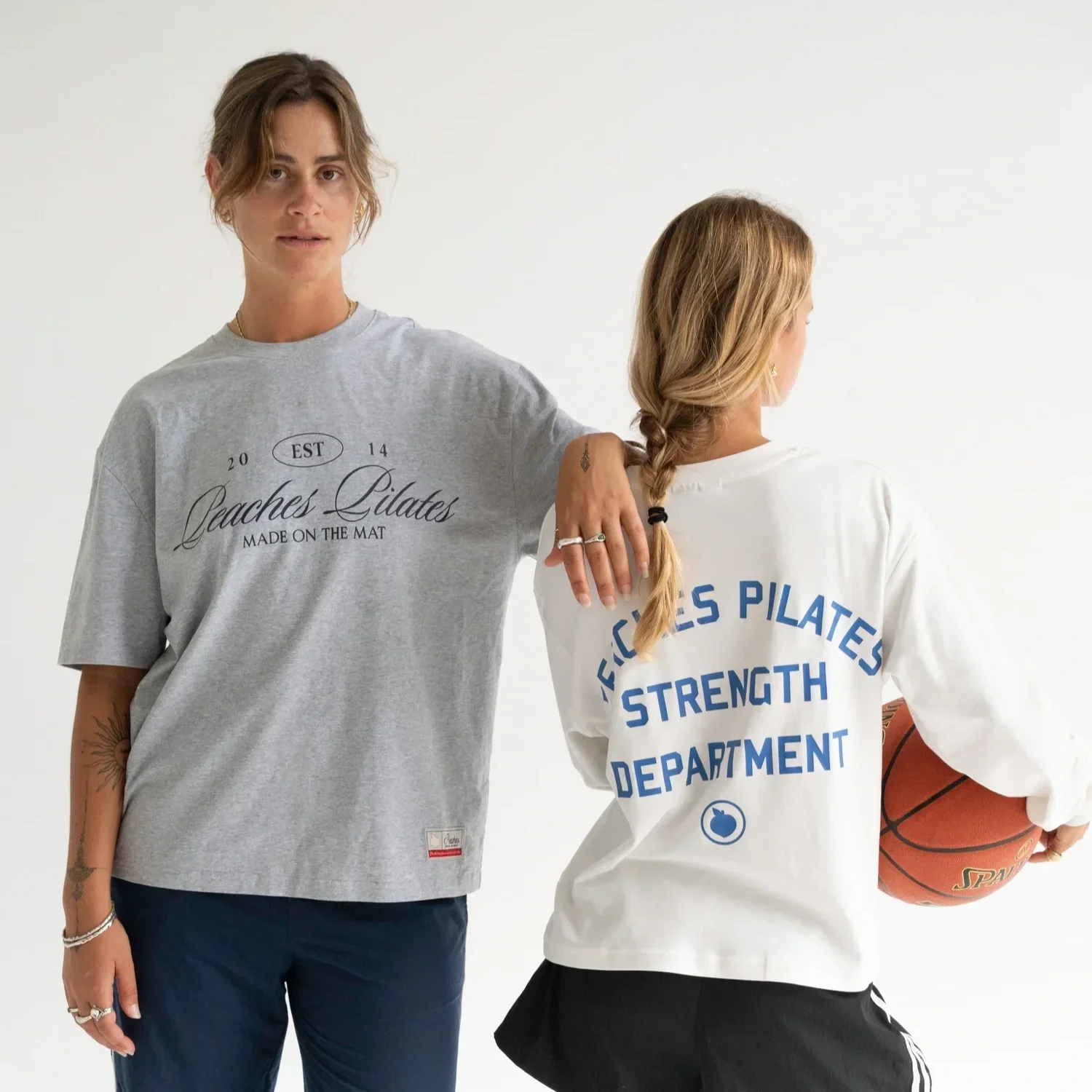 A woman on the left wearing a gray t-shirt with the text 'Peaches Lulates Made on the Mat' and dark pants. A woman on the right, partially visible, wearing a white t-shirt with blue text and holding a basketball, with her back turned.