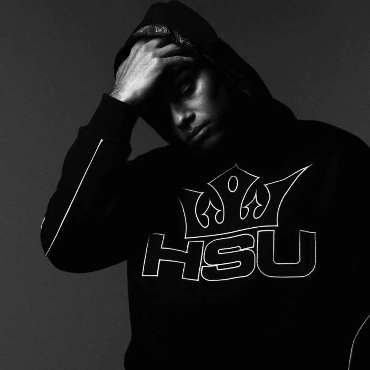 A young man wearing a hoodie with the hood up, with his hand on his forehead, looking down, in black and white.