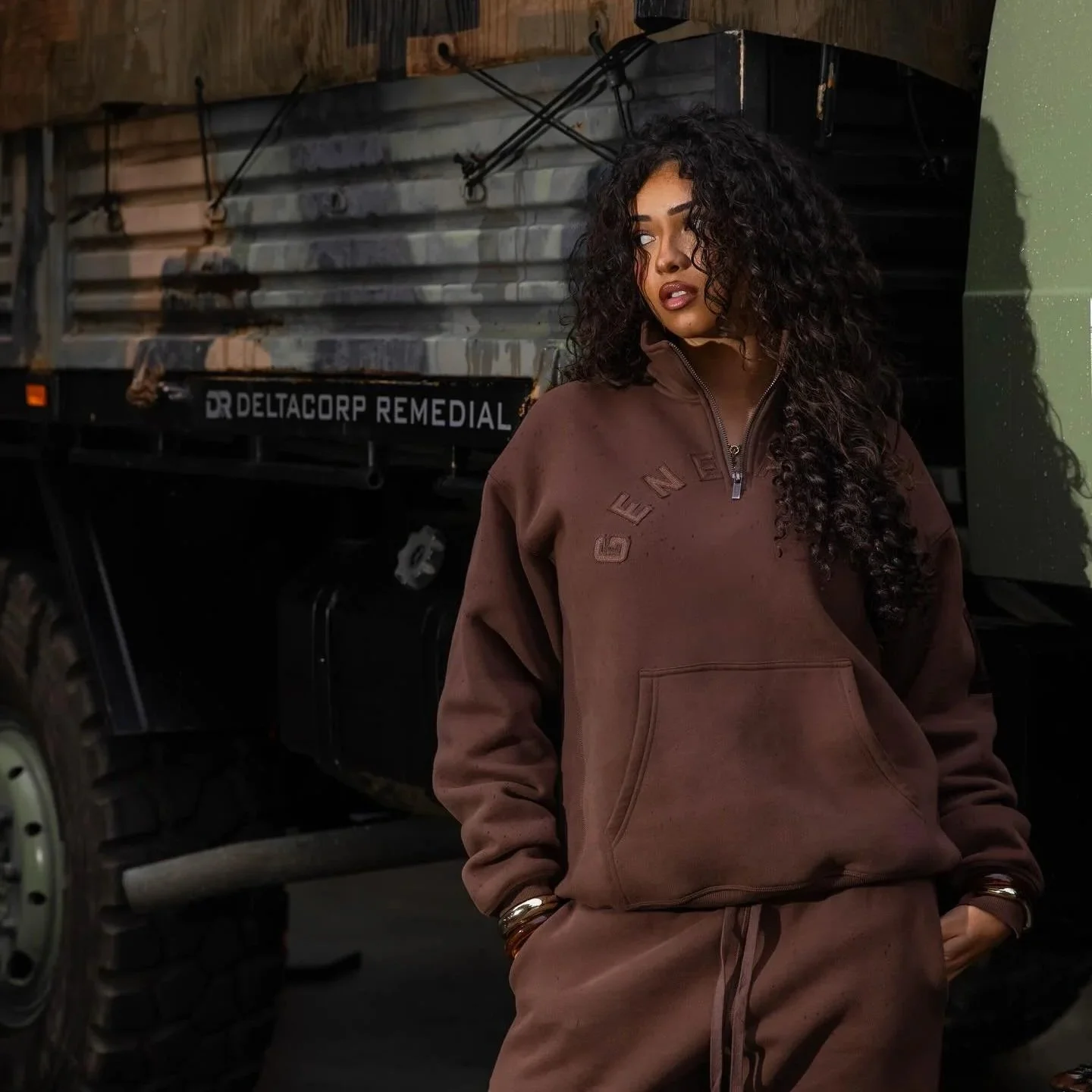 A young woman with curly black hair wearing a brown sweatshirt and matching sweatpants standing in front of a vehicle with a large off-road tire and a black cargo box. She has a bracelet on her wrist and her hands are in her pockets.