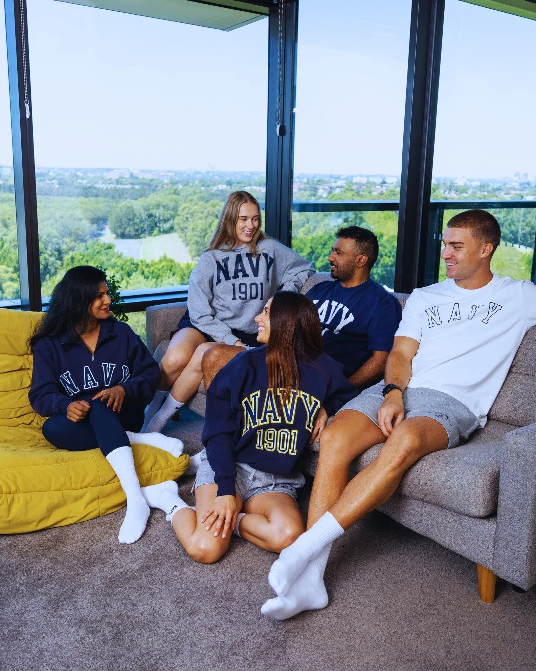 A group of five young adults with diverse ethnicities wearing Navy sportswear, sitting and standing on a gray sofa and yellow bean bag in a modern apartment with large windows overlooking a green park and city skyline.