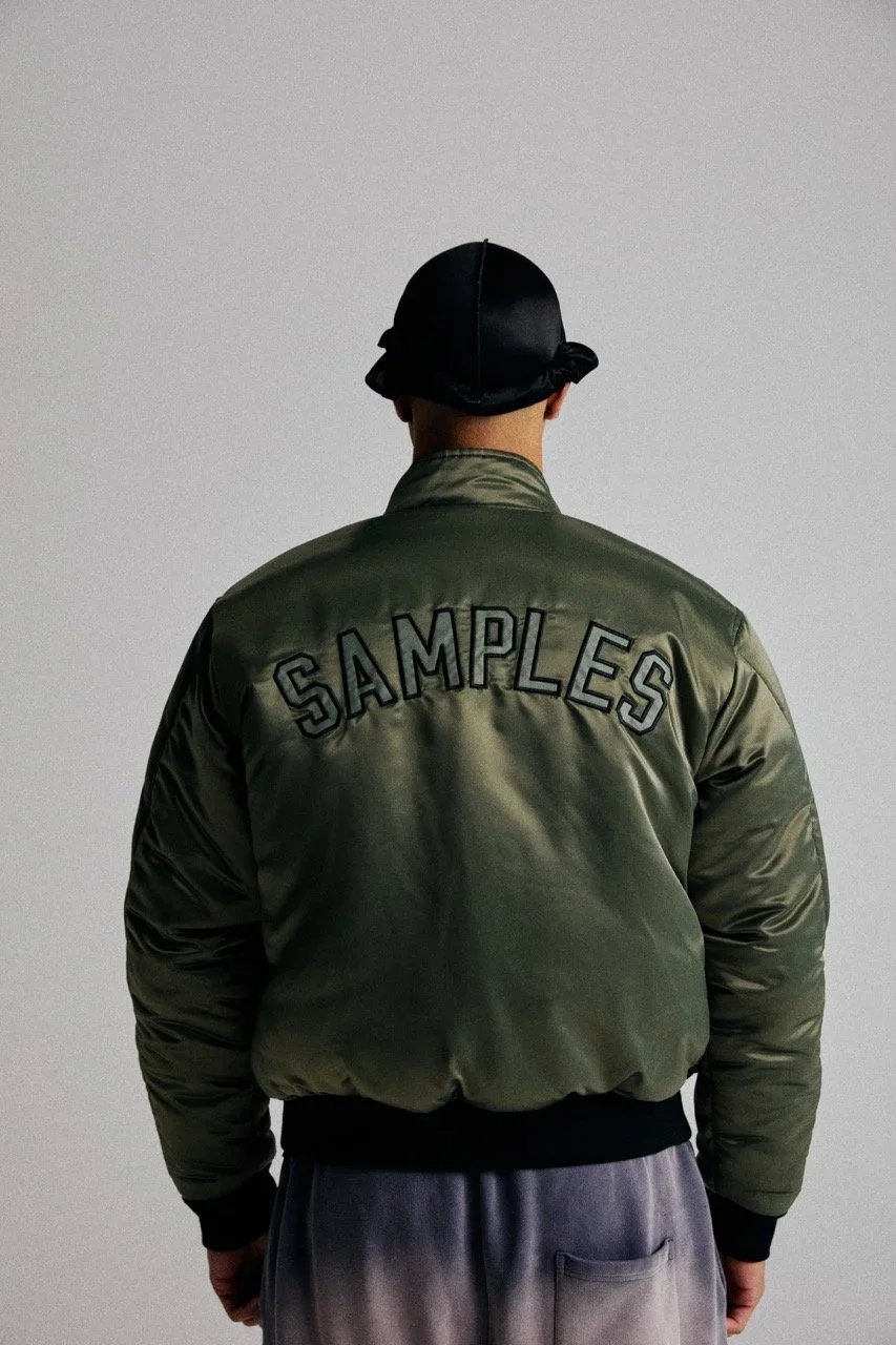 Back view of a person wearing an olive green bomber jacket with the word 'SAMPLES' embroidered on the back, paired with gray pants and a black head covering.