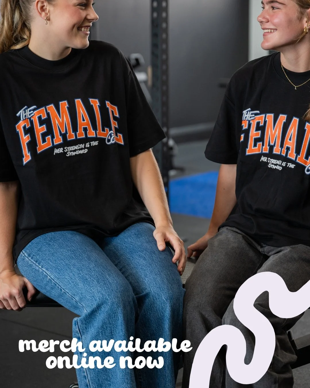 Two women sitting and smiling at each other, wearing black T-shirts with the text "The Female" in orange and blue, and smaller text "Her strength is the standard." The background appears to be a gym or training room. Overlaid text reads "merch available online now."