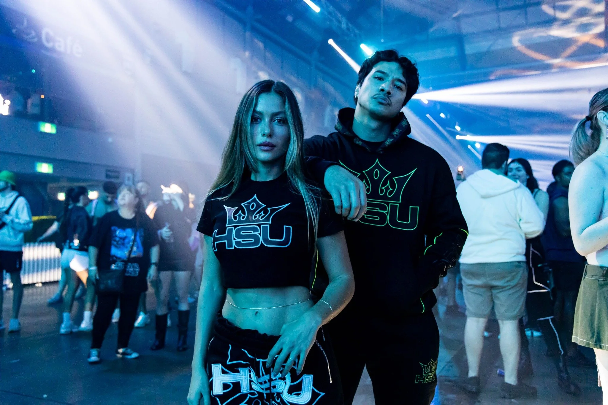 Two young adults, a woman with long blonde hair and a man with dark hair, wearing black clothing with neon green and blue designs, posing together at a nightclub with blue lighting and other people dancing in the background.