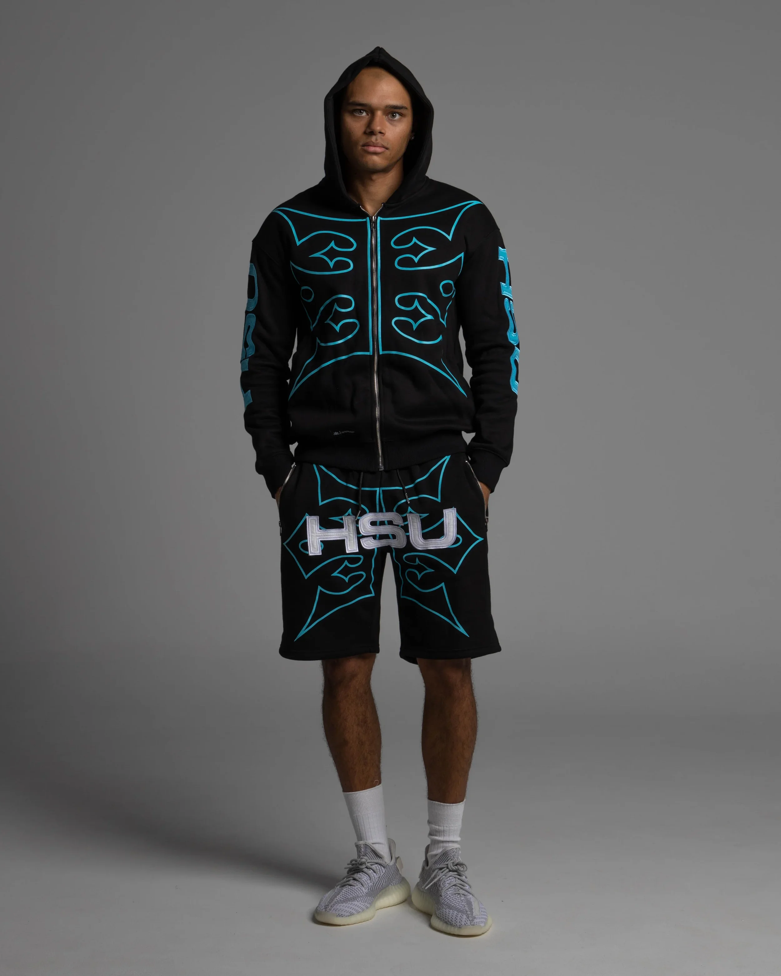 A young man standing against a gray background, wearing a black hoodie with blue outlines and designs, matching black shorts with blue outlines, white sneakers, and white socks.