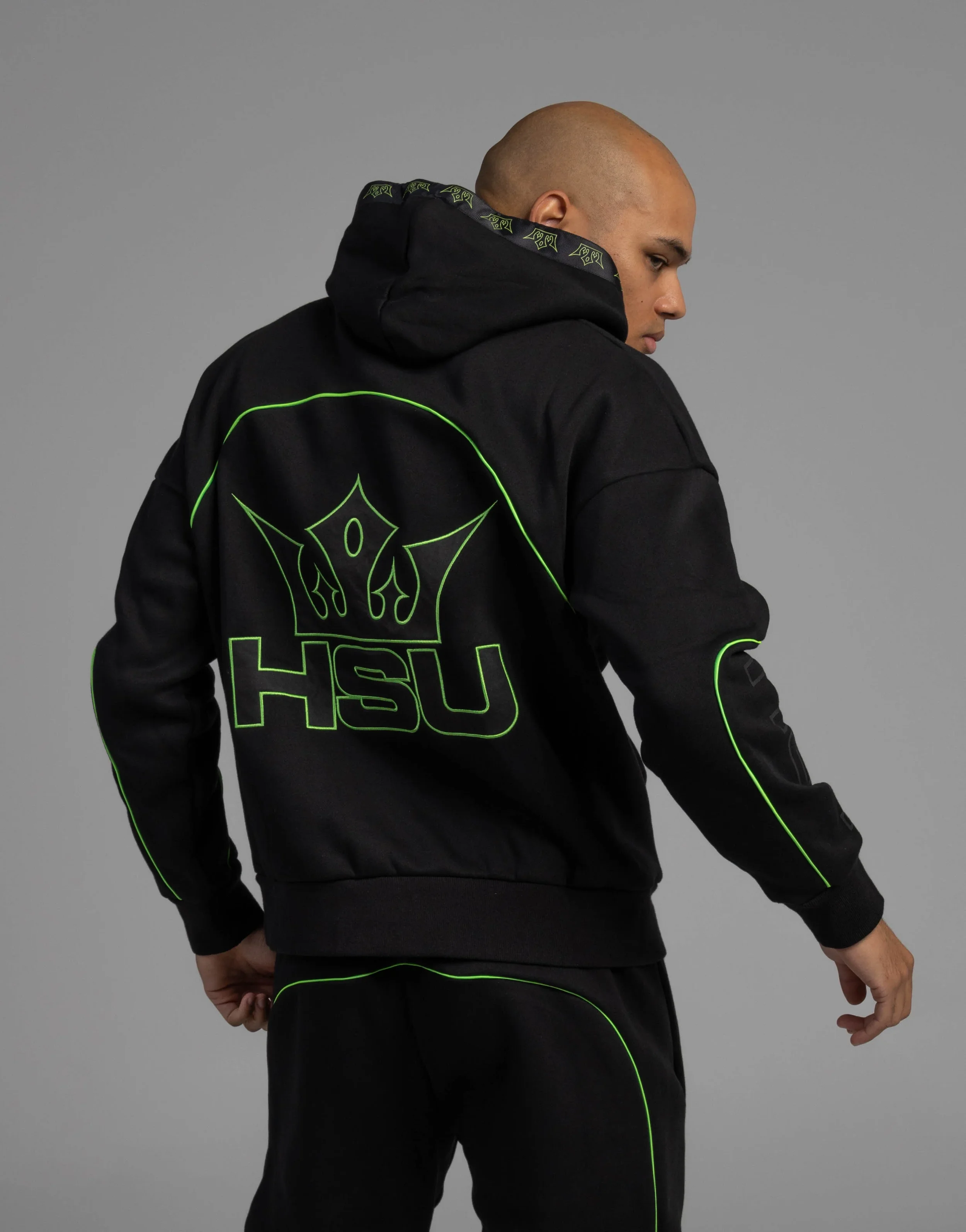 A person with a shaved head wearing a black hoodie with neon green outlines and designs, standing against a plain gray background. The person is turned to the side, looking down, with one hand resting on their hip and the other hanging down.