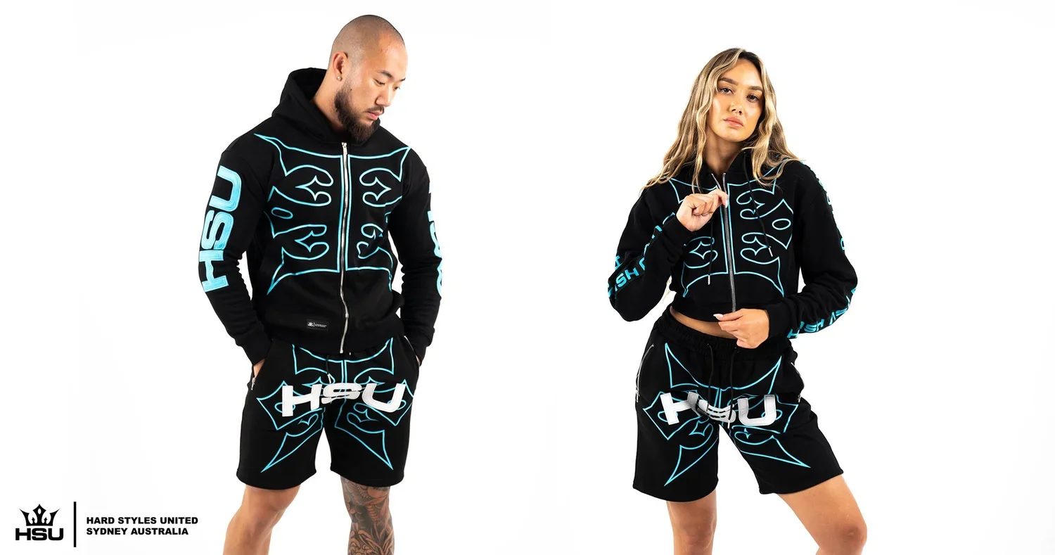 Two models wearing matching black hoodies and shorts with blue and white graphic designs and the HSU logo. The male model is looking down with hands in his pockets, and the female model is looking forward holding the hoodie zipper.