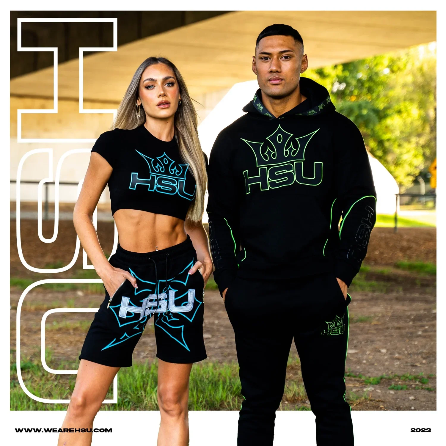 A man and woman wearing black athletic clothing with HSU logos, standing outdoors with trees and a structure in the background.
