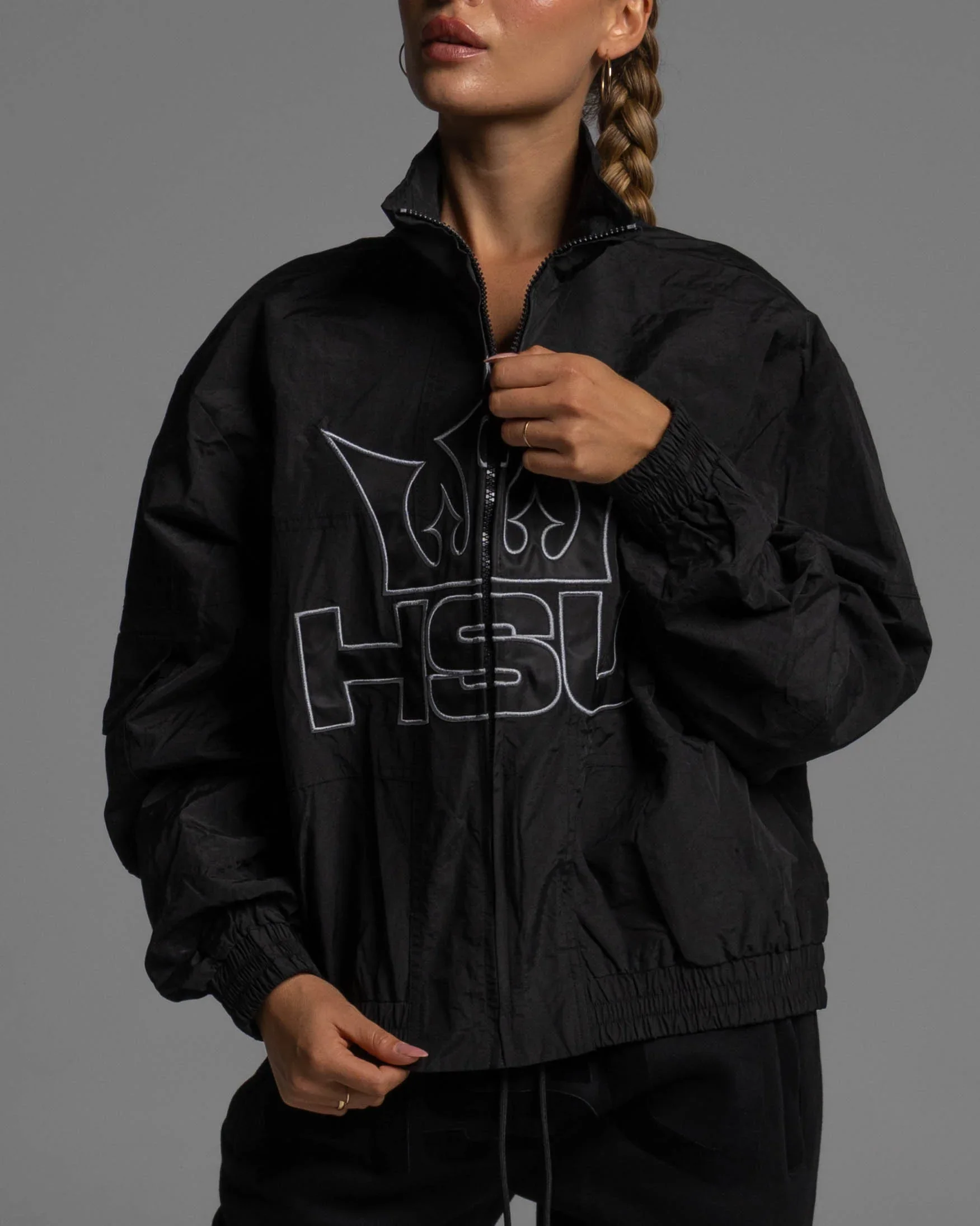 A woman wearing a black North Face jacket with white outline logos, holding the zipper with one hand, standing against a gray background.