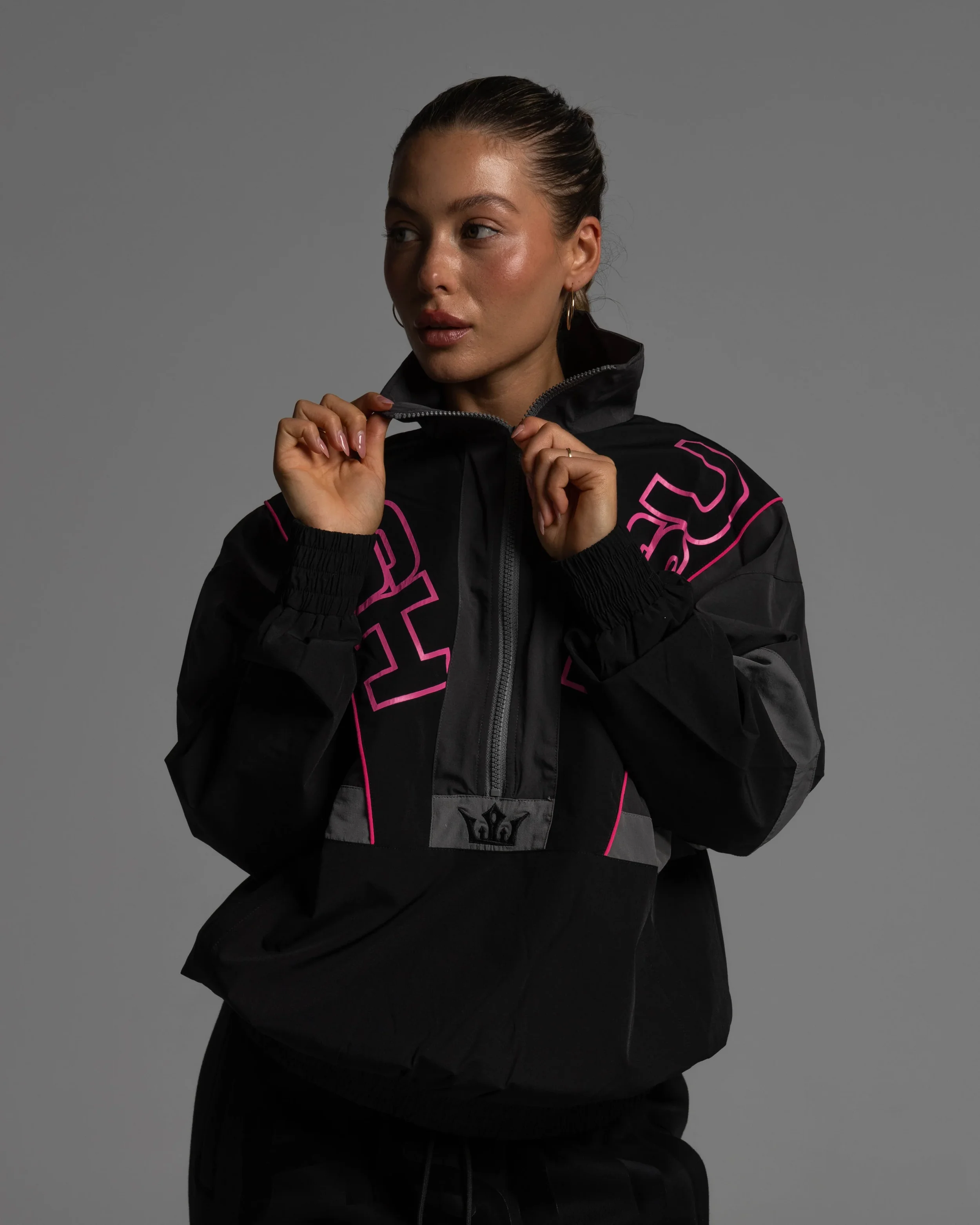A woman with slicked-back hair wearing a black zip-up jacket with pink lettering, holding the collar with both hands, against a plain gray background.