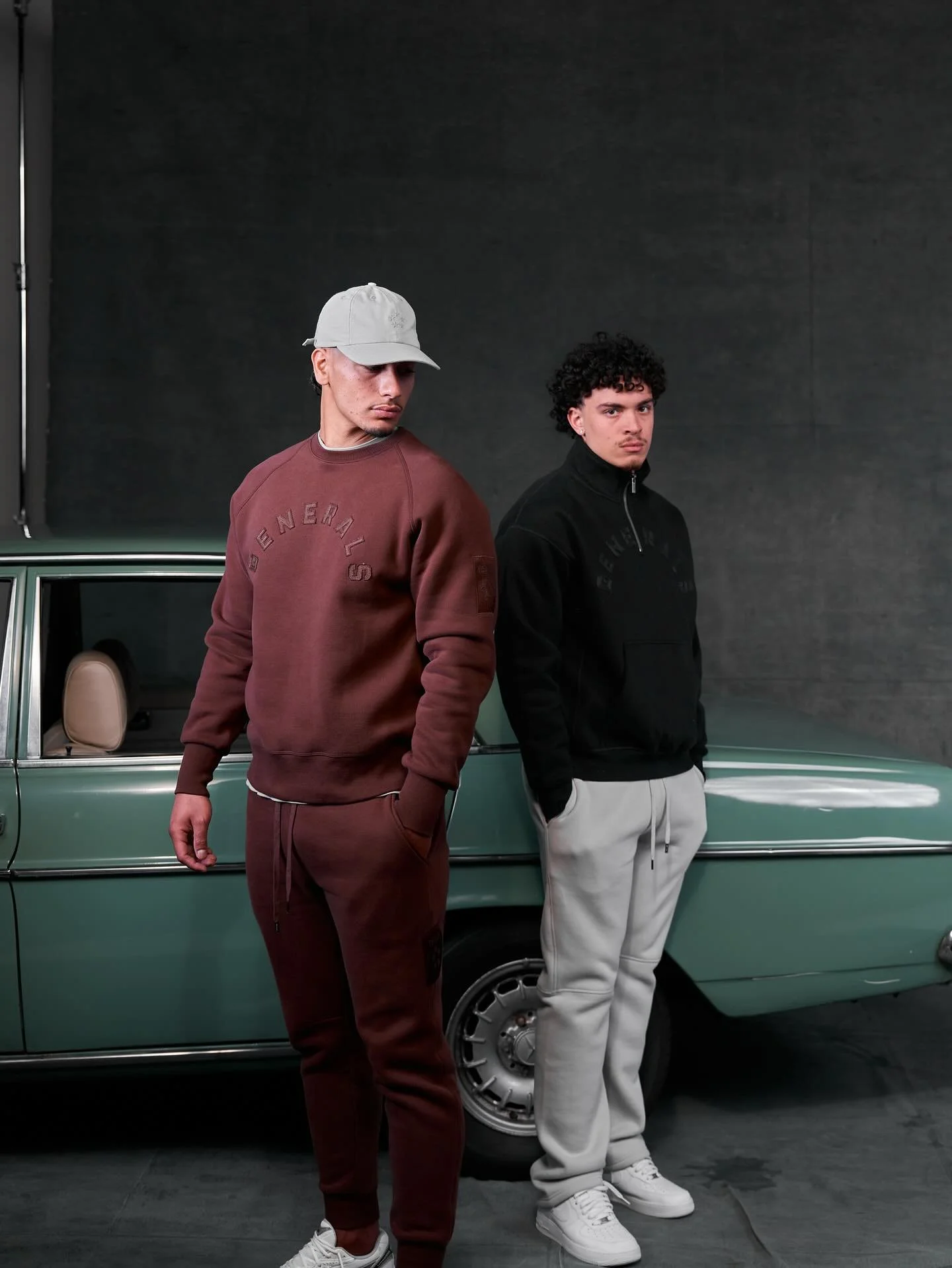Two young men standing in front of a green vintage car against a plain dark wall. The man on the left wears a beige cap, maroon sweatshirt and sweatpants, while the man on the right wears a black zip-up jacket and beige sweatpants. Both have casual sneakers and serious expressions.