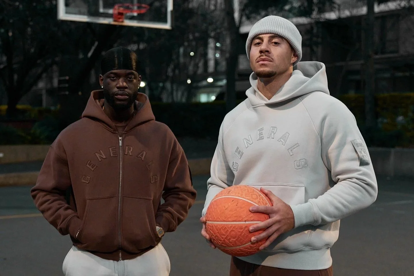 Two men standing on a basketball court at dusk, one holding an orange basketball and wearing a white hoodie, the other in a brown hoodie.