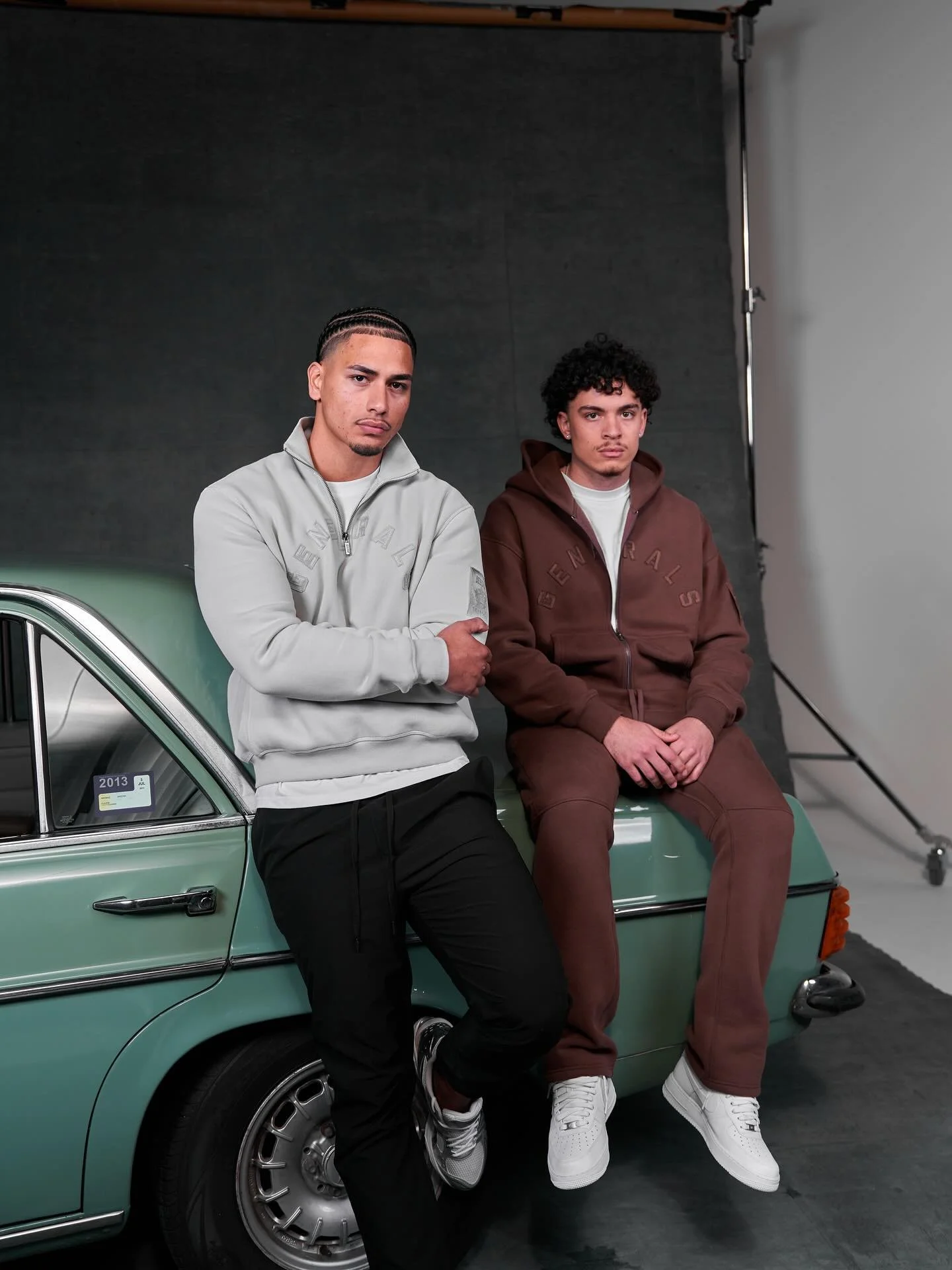 Two young men sitting on the hood of a vintage green car in a photo studio. One man in a light gray jacket and black pants stands with arms crossed, and the other in a brown hoodie and brown pants sits with hands clasped.
