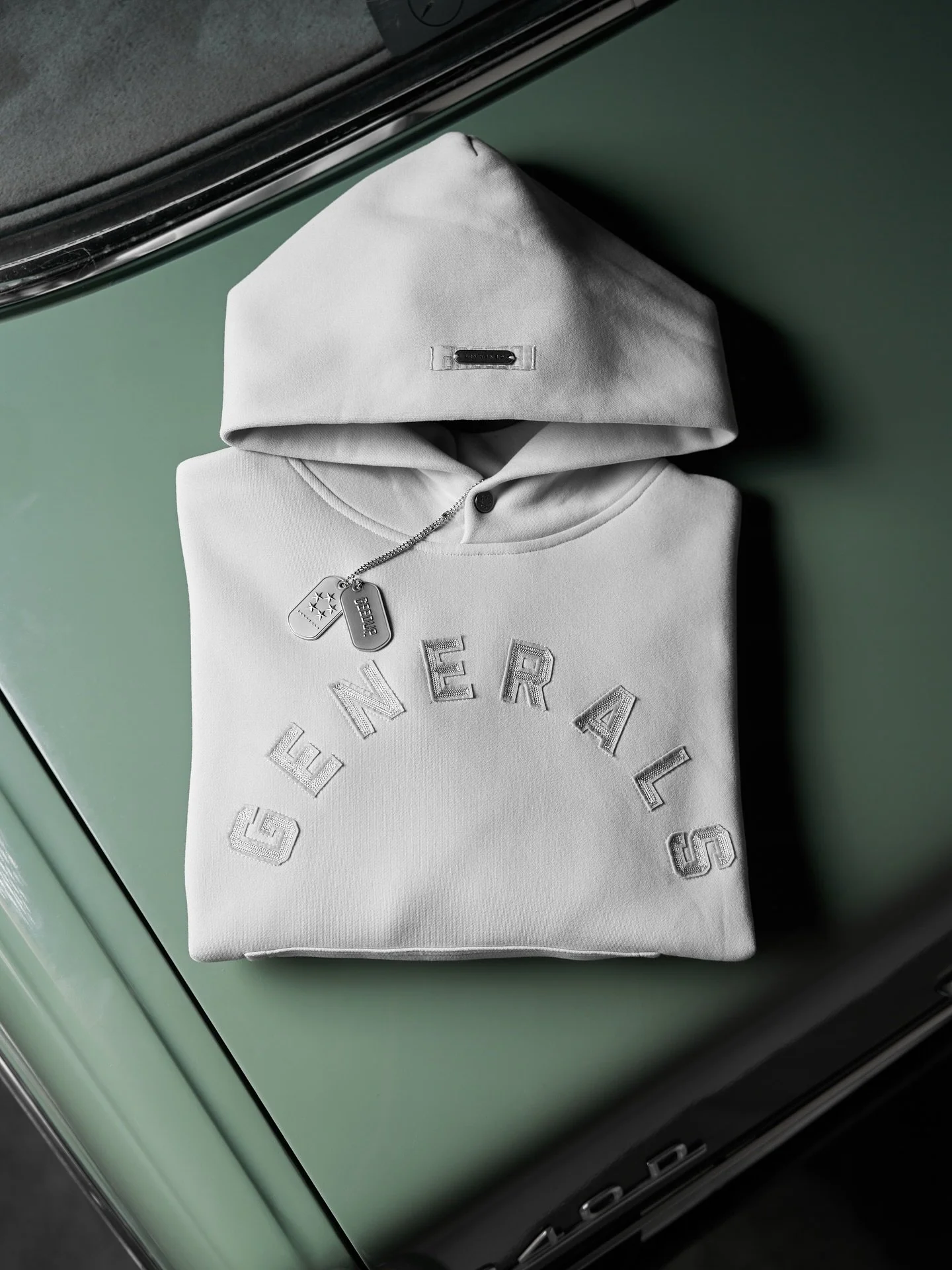 A folded white sweatshirt with initials and embroidered text, with a silver chain necklace with a dog tag and stars, placed on a reflective green surface.