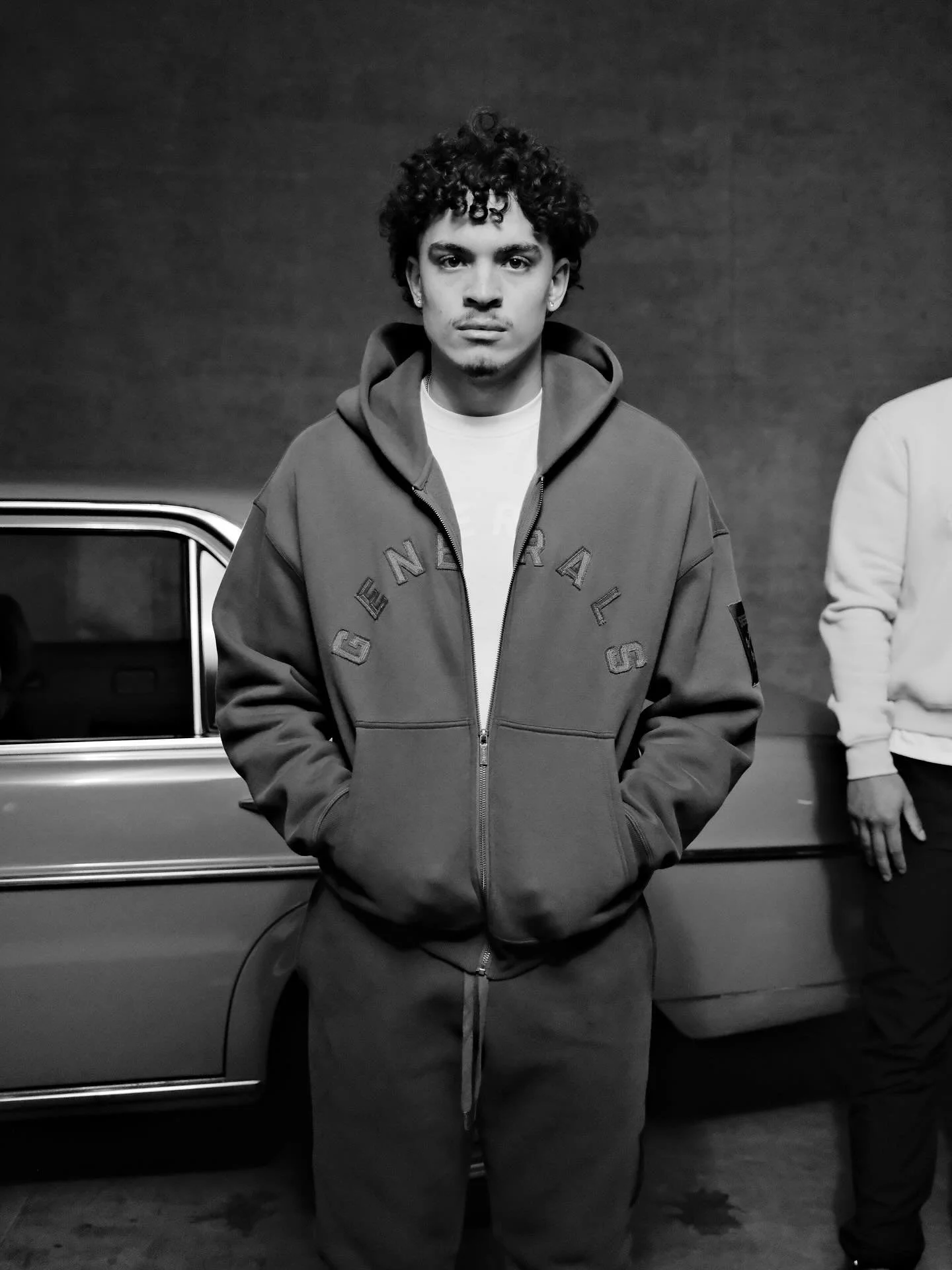 A young man with curly hair, wearing a hoodie and sweatpants, standing in front of a car in a garage or parking area. Another person is partially visible to the right.