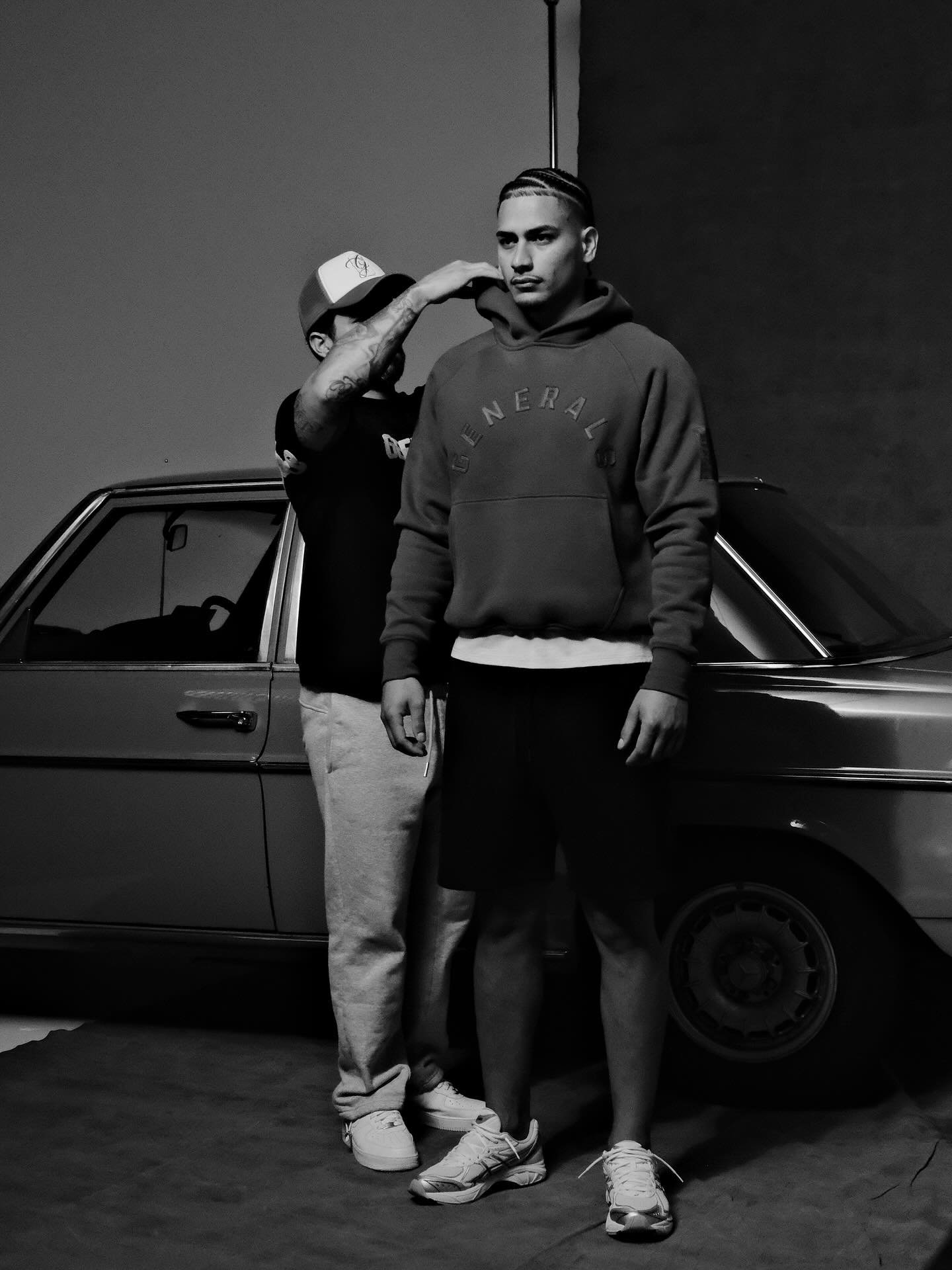 Two young men, one wearing a hoodie and shorts, and the other in sweatpants and sneakers, stand close together in front of a car in a dark environment. One man is adjusting the other's collar.