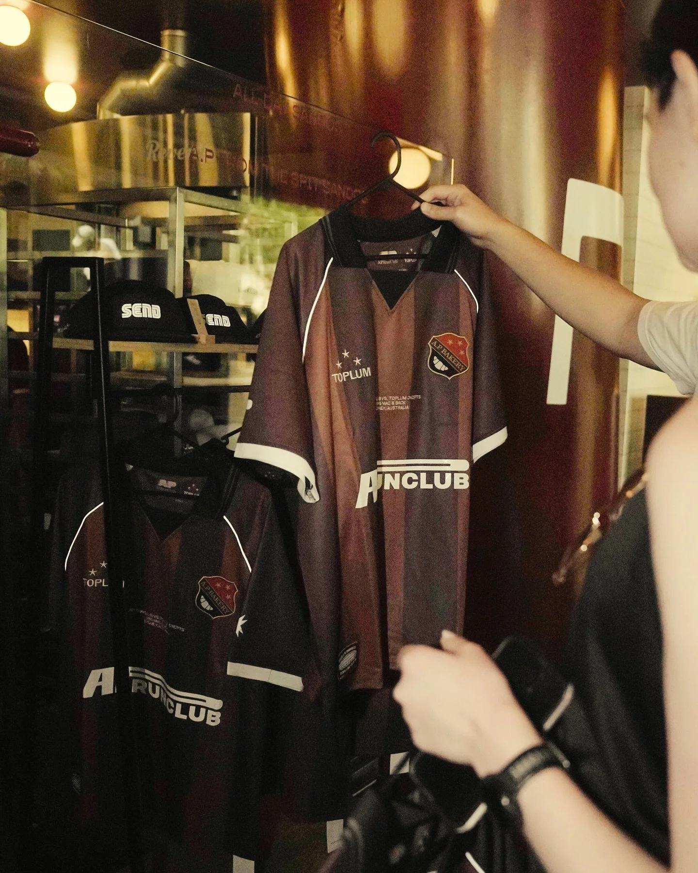 A person is holding up a maroon and black sports jersey with a shield emblem and the words "AFL Bakery" and "Toplum" printed on it, in a retail store. The store has a display shelf with more jerseys and black caps, and a reflective glass window behind the display.