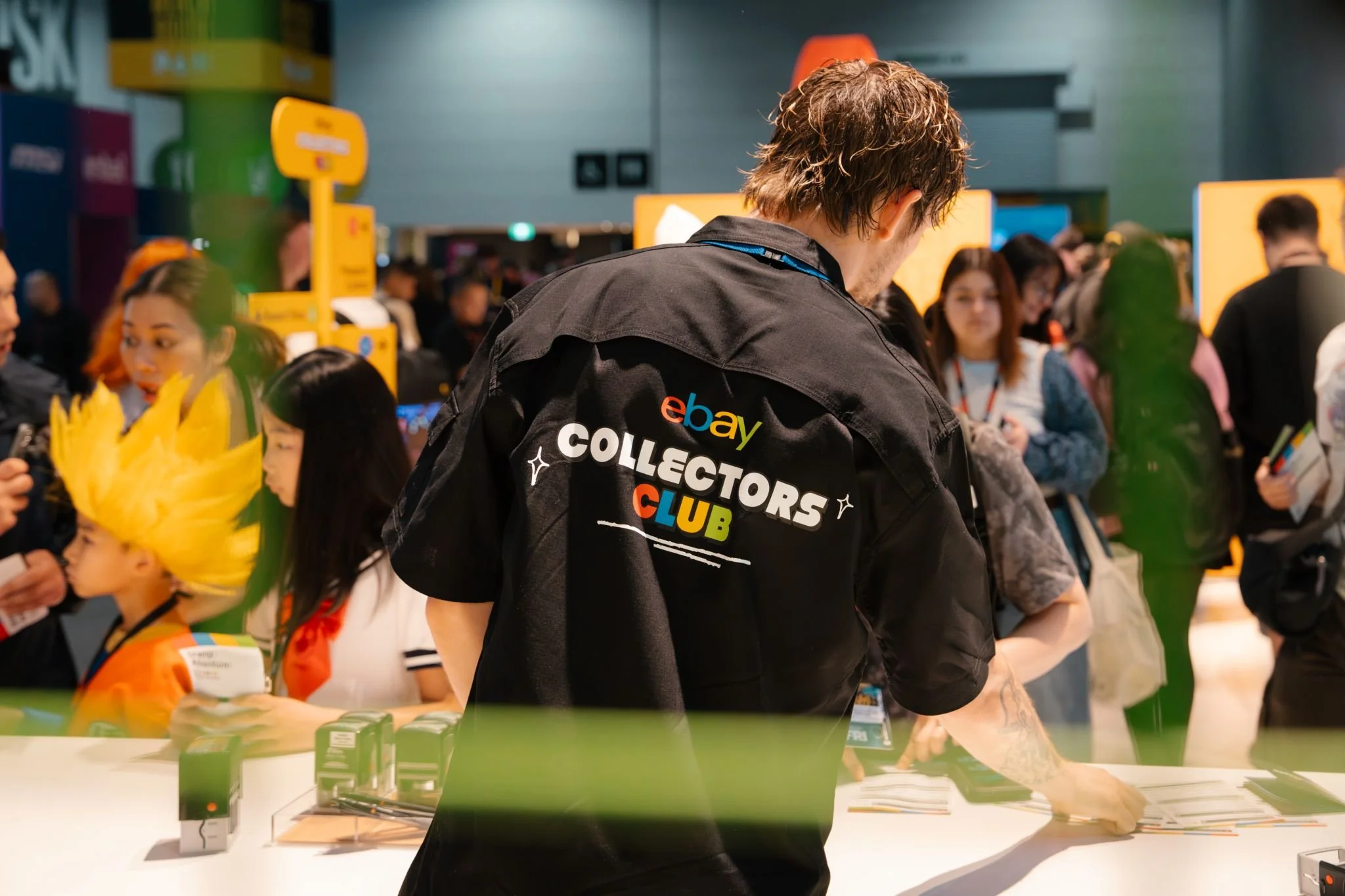 A man wearing a black eBay Collectors Club jacket is at a booth or table with several small boxed items. There are Chinese lanterns and a crowd of people, some with colorful hair, in the background at an indoor event or convention.