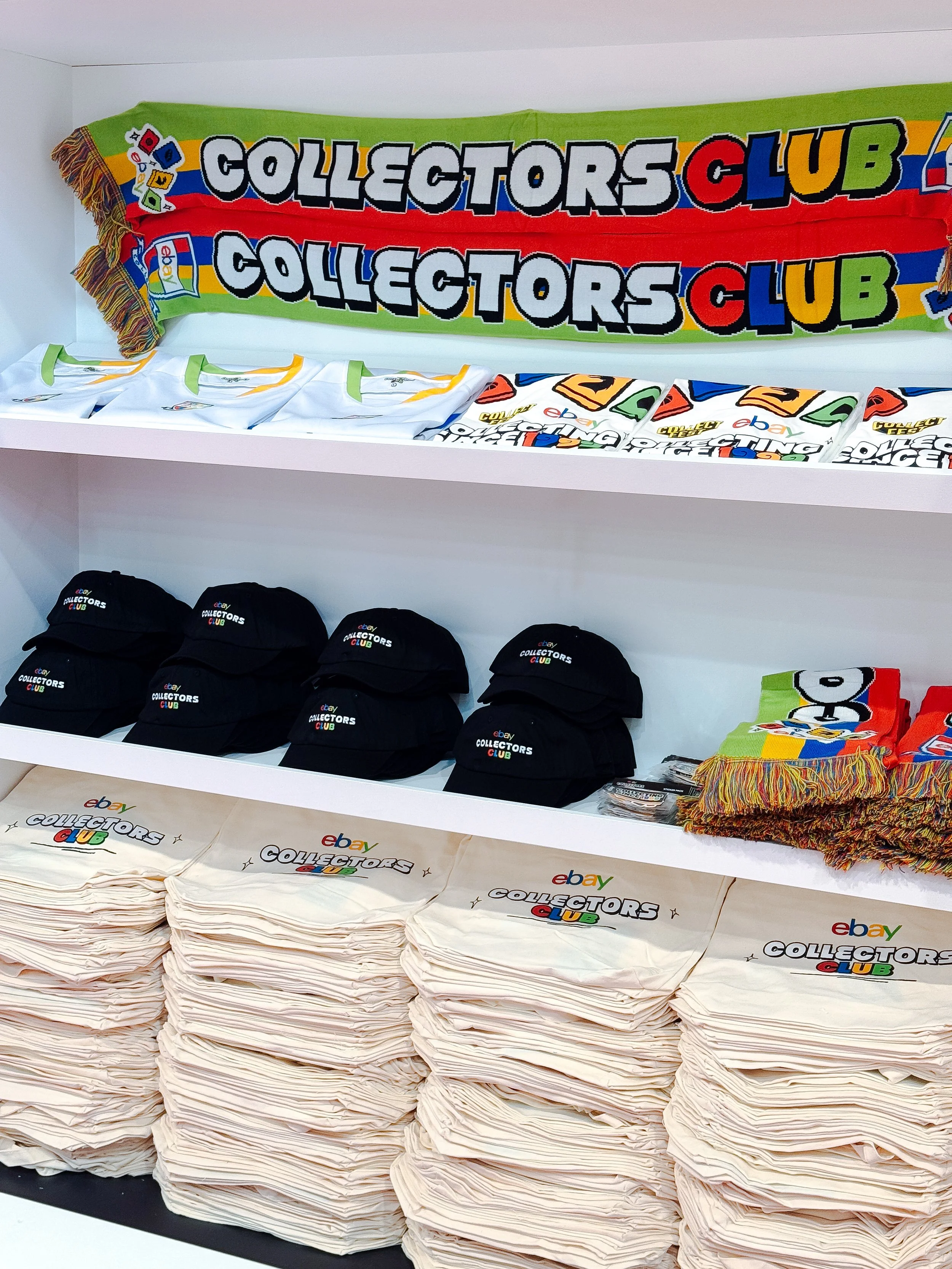 Display of eBay Collectors Club merchandise, including t-shirts, hats, scarves, and scarves with the Collectors Club logo, arranged on shelves.