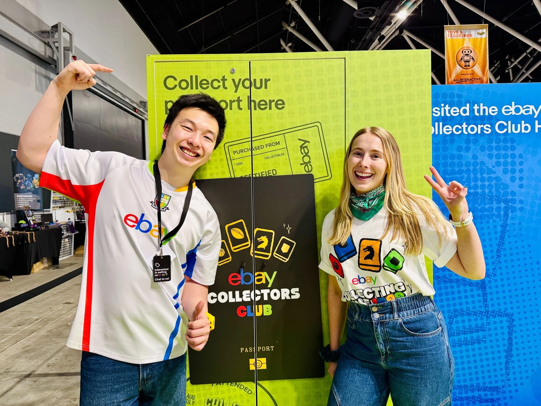 Two smiling young people making peace signs in front of a large green and blue sign promoting an eBay collectors club at a convention or event.
