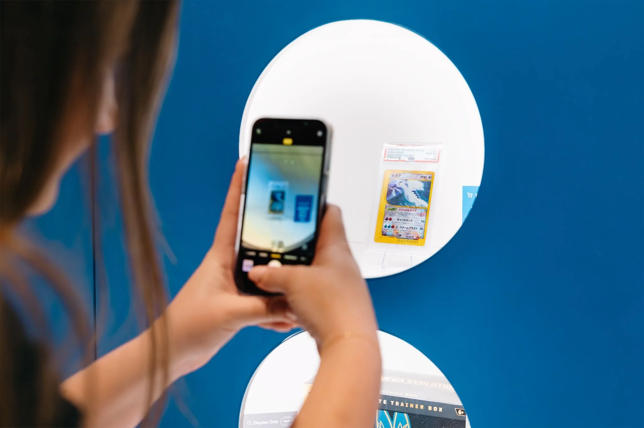 Person taking a photo of a Pokémon card reflected in a circular mirror, with a Pokémon card featuring Lugia visible.